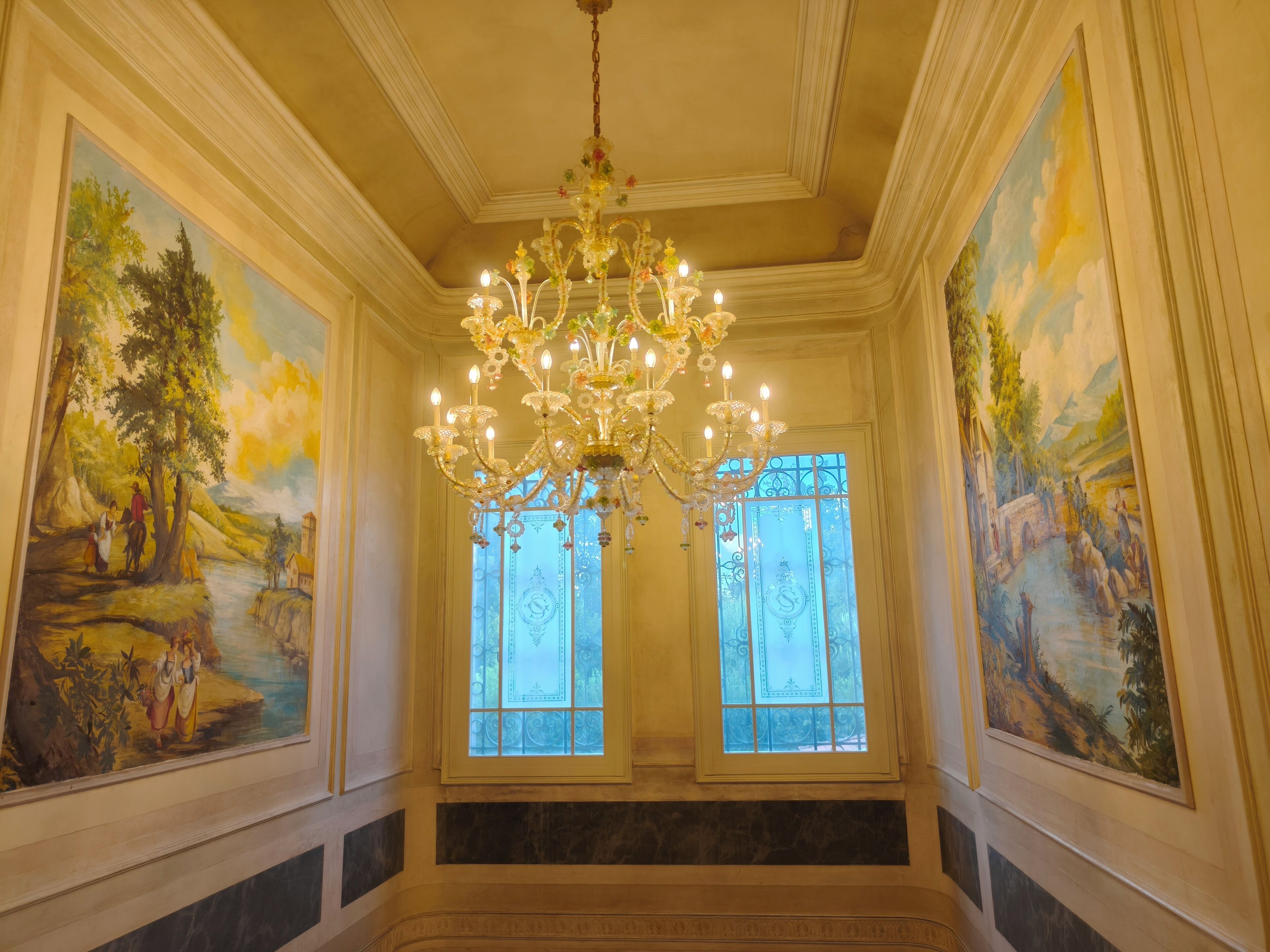 Un salón imponente con candelabro, ventanas amplias y pinturas murales de estilo clásico.