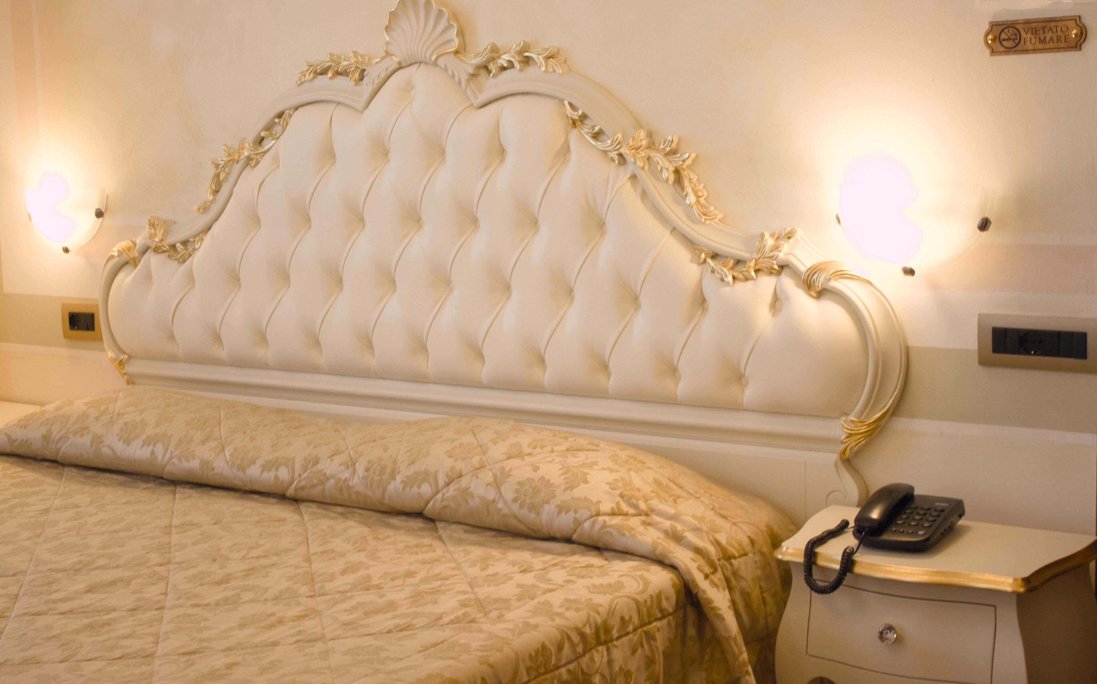 A hotel room with a tufted headboard, a bed with a patterned bedspread, a bedside table with a telephone and a lamp, and a wall-mounted light fixture.