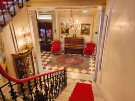 A grand staircase with a red carpet leading to a room with a chandelier, red chairs, and a wooden cabinet.