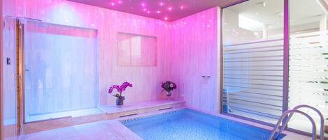 A modern indoor swimming pool with a glass wall, pink ambient lighting, and a small bench with flowers.