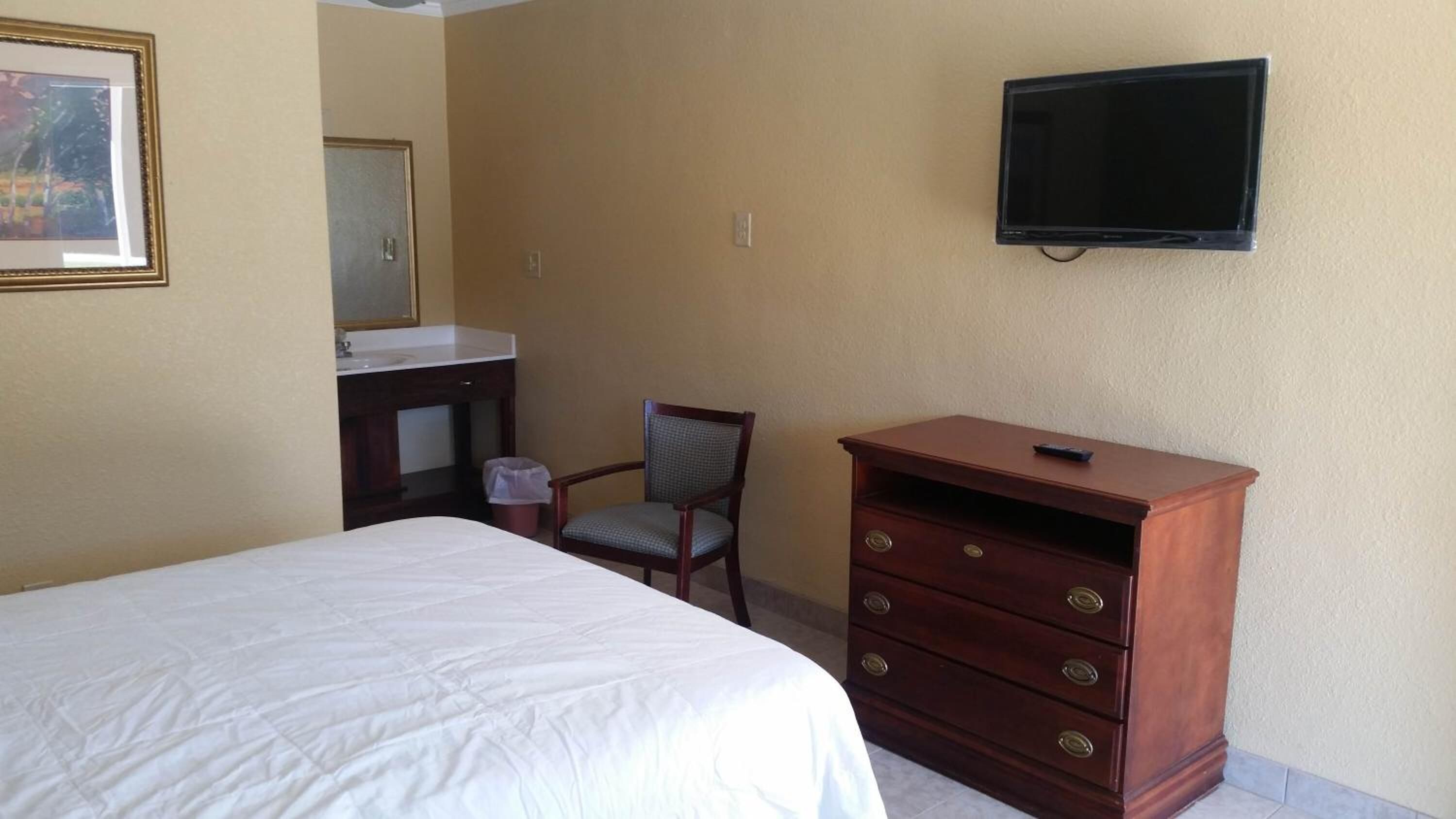 Habitación de hotel con cama, cómoda de madera, silla, mesita, televisor montado en la pared y baño con lavabo y espejo.