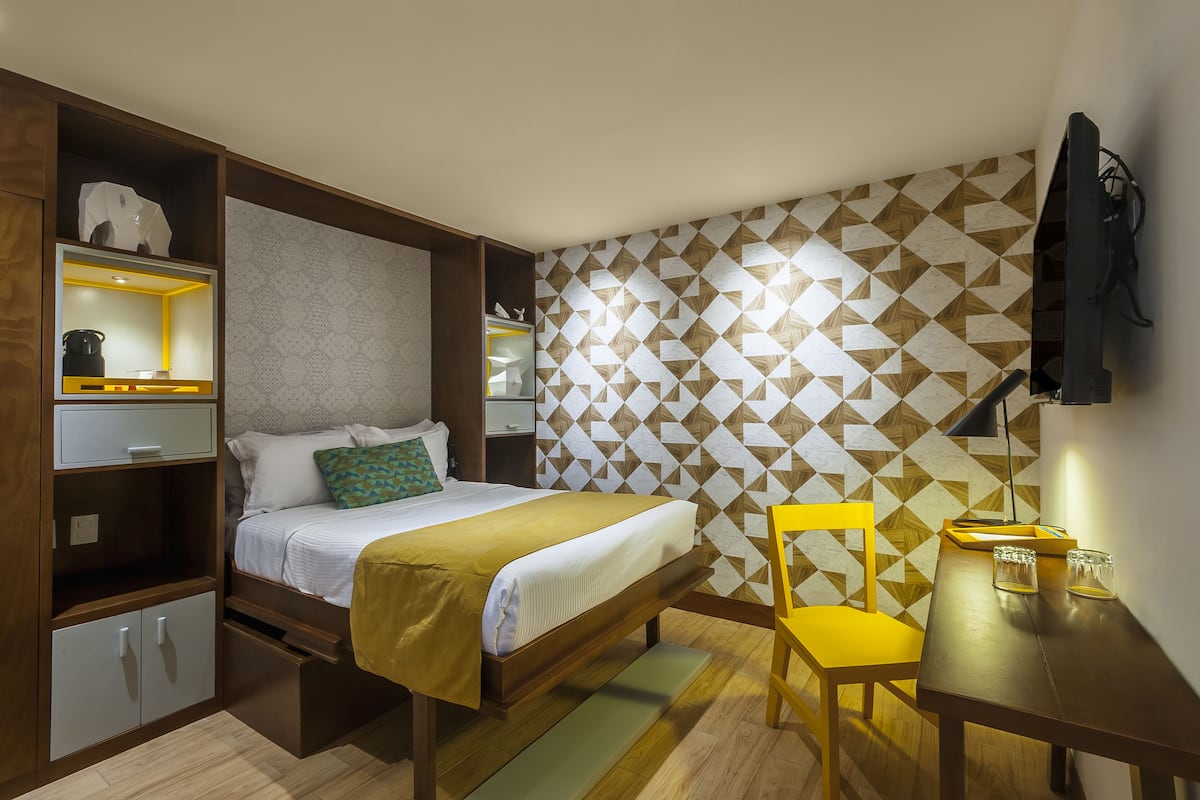 Habitación de hotel compacta con cama, escritorio, silla, televisor y pared con diseño.