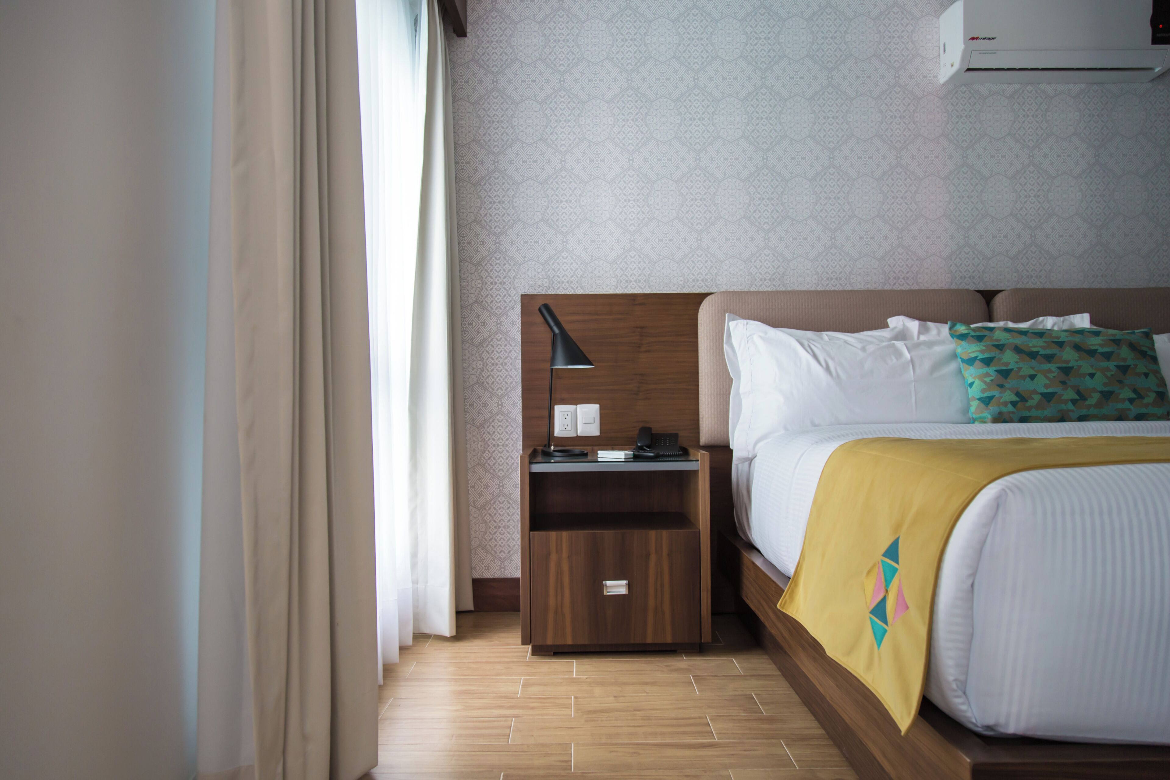 Habitación de hotel con cabecera de madera, una mesita de noche con lámpara, teléfono y un cajón pequeño.