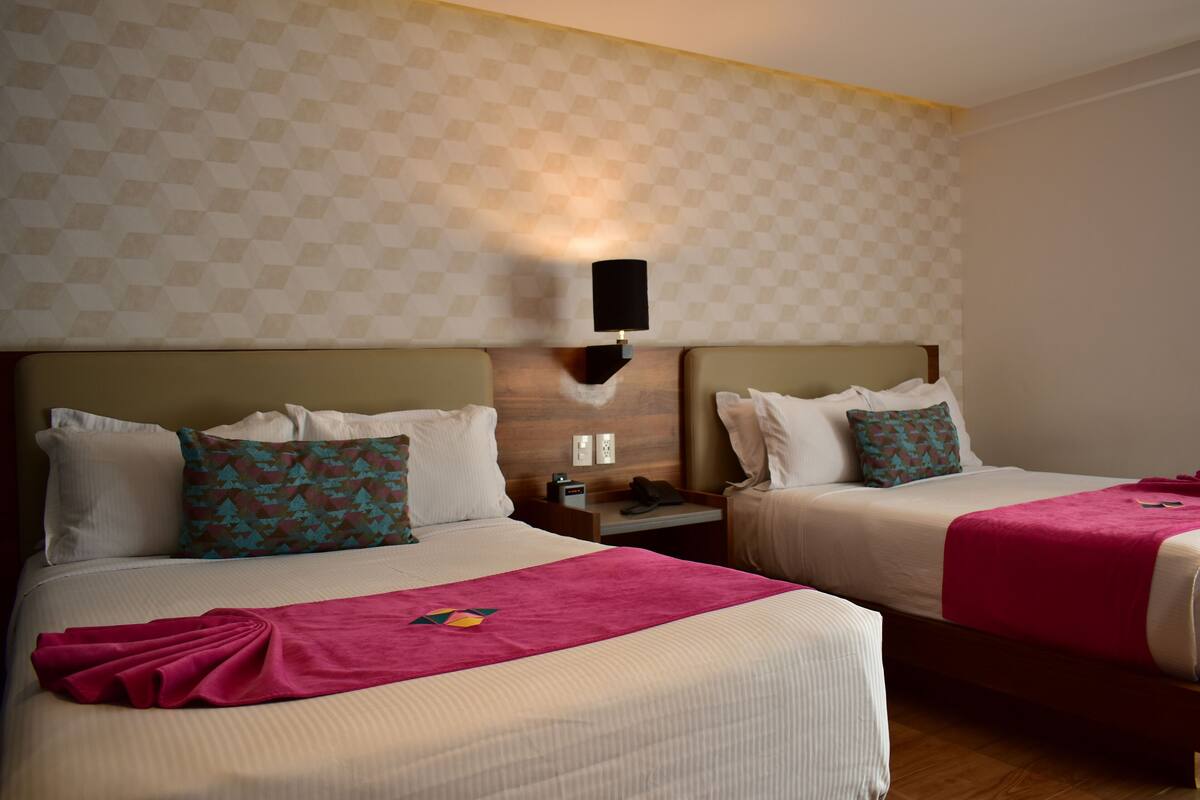 Habitación de hotel con dos camas, cada una con ropa de cama blanca y almohadas decorativas. Hay una lámpara de pared sobre las camas, y una mesita con un teléfono y un libro.