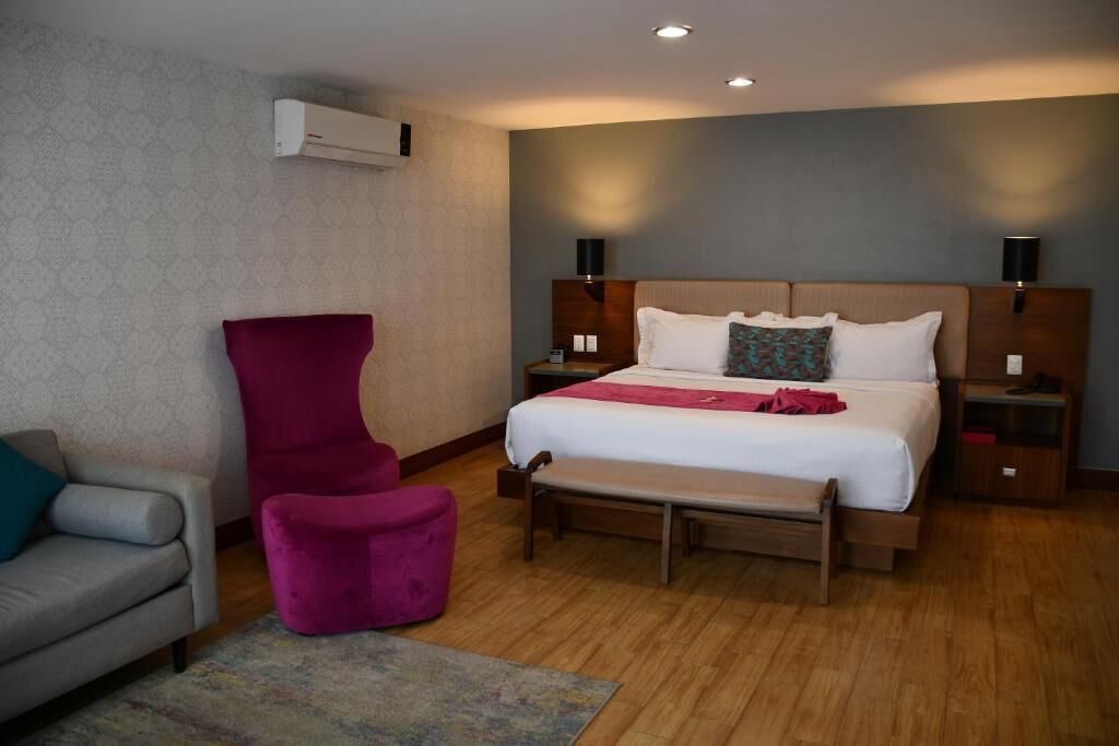 Een hotelkamer met een bed, een stoel, een bankje, een nachtkastje en een airconditioner.