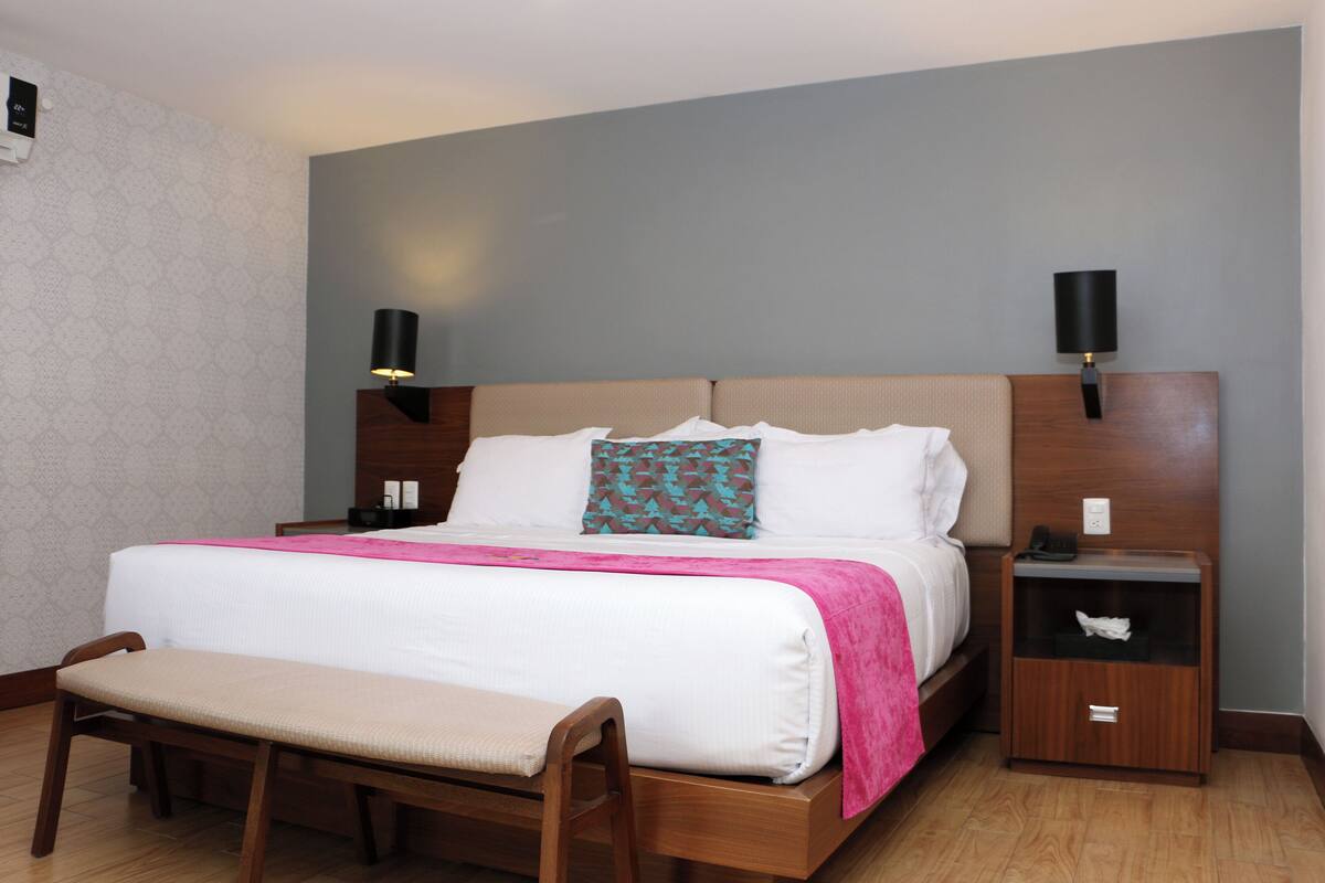 Una cama bien hecha con edredón blanco y manta rosa, una mesita de noche de madera con una lámpara, y un banco al pie de la cama.