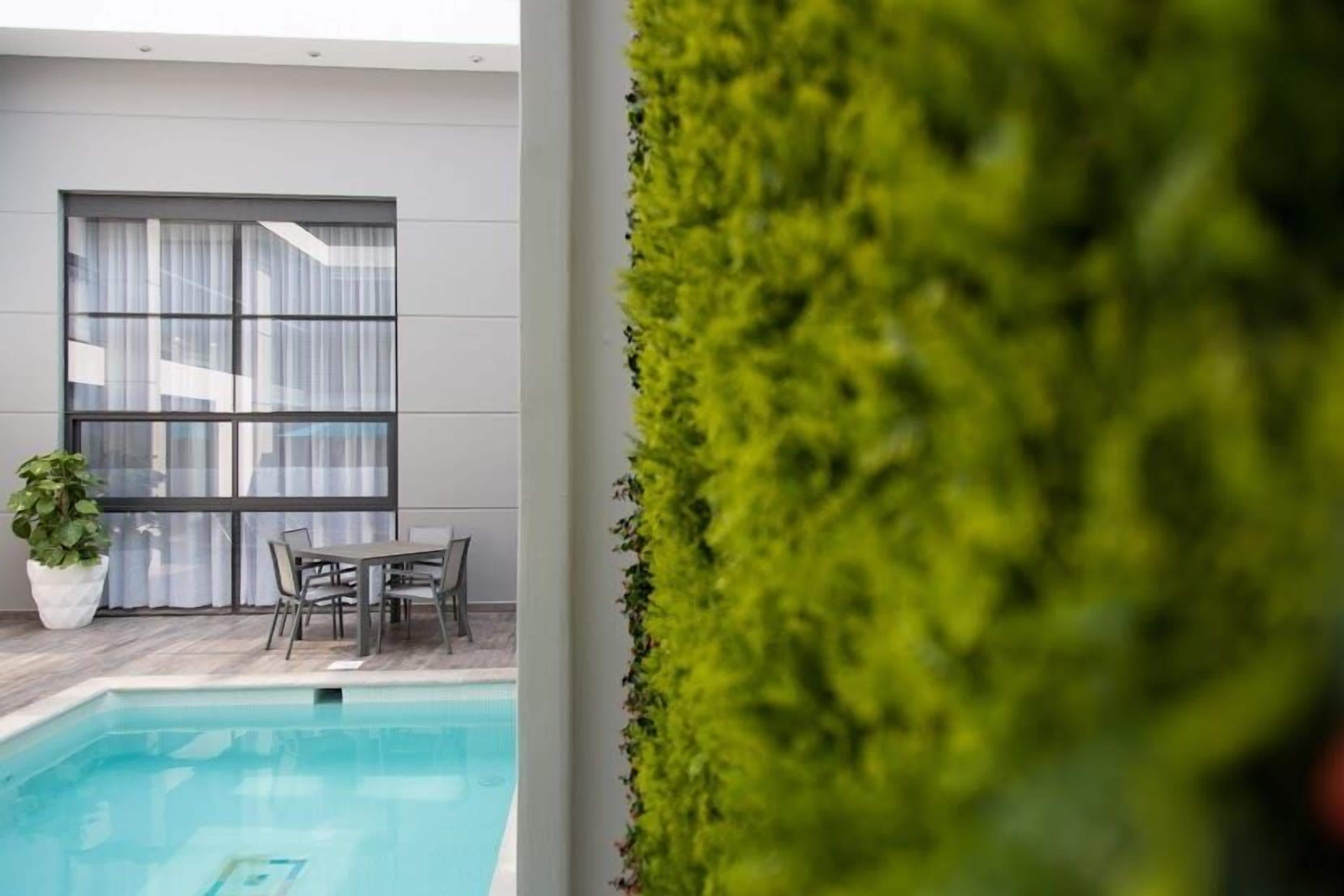 Zona de piscina con una mesa y sillas, una planta en maceta y vistas a un edificio con grandes ventanales.