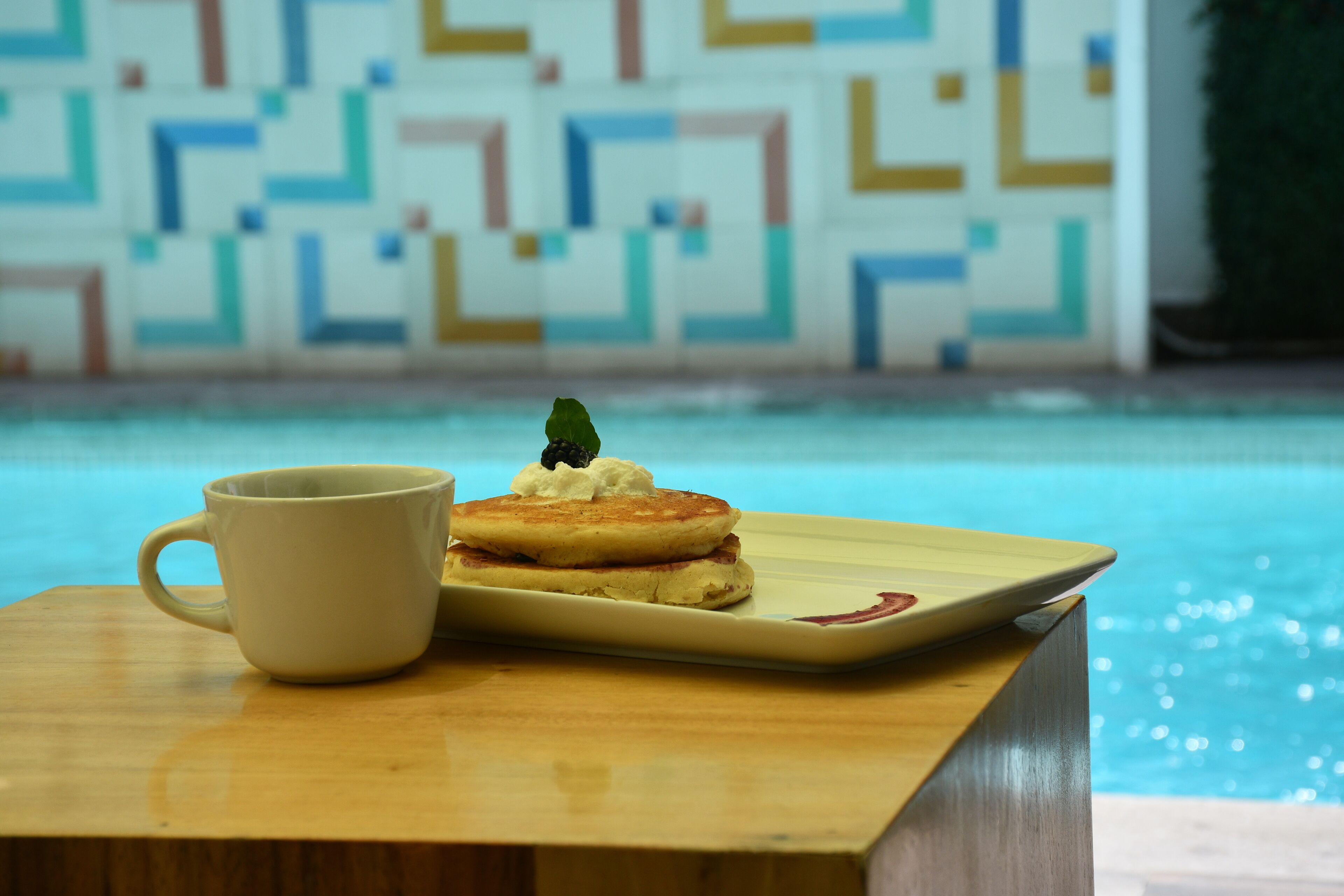 Ett träbord med en vit mugg och pannkakor med vispad grädde och ett körsbär, serverat på en fyrkantig tallrik, nära en pool med en vägg som är klädd med geometriska plattor.
