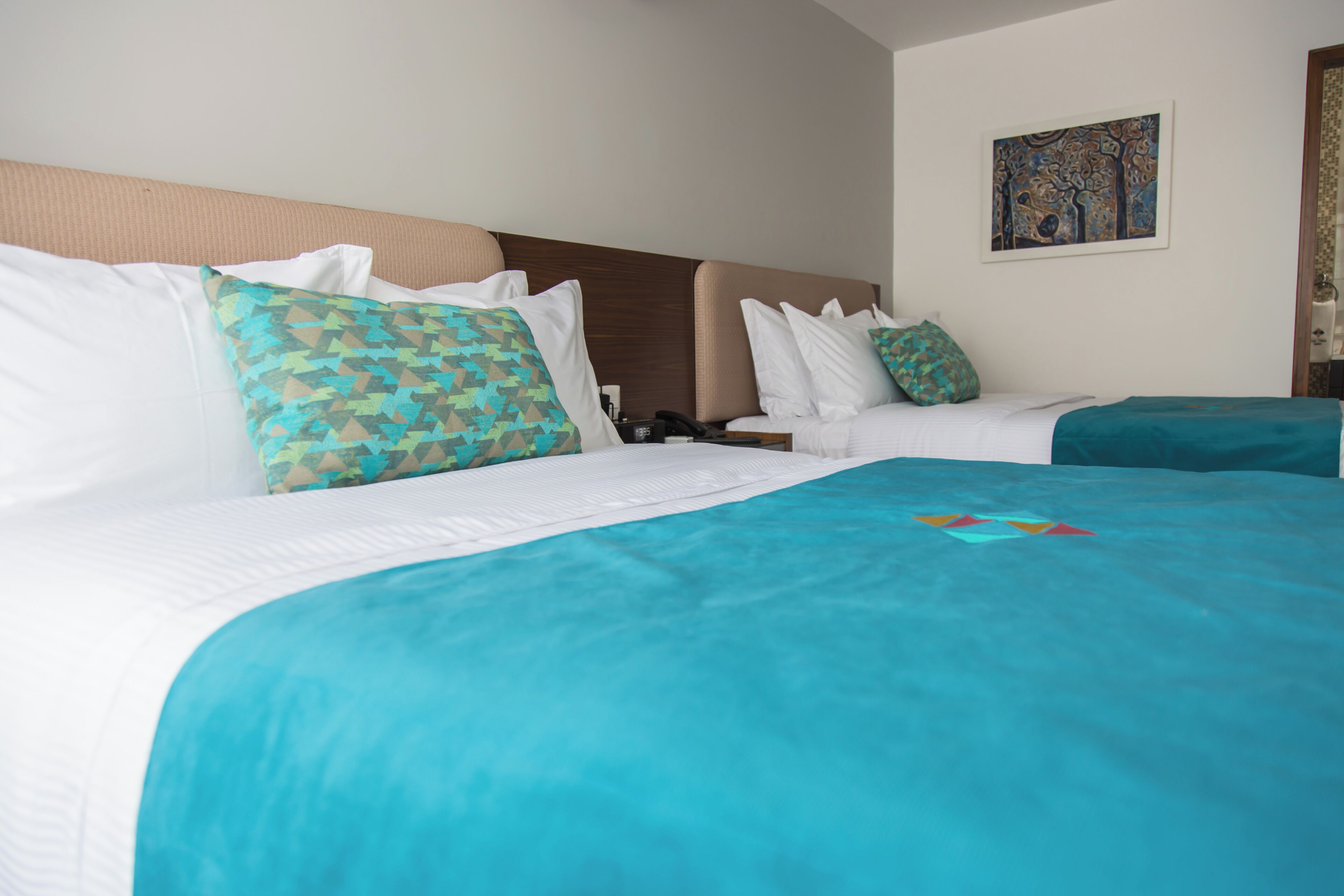 A hotel room with two beds, each with a patterned pillow and a teal bedspread.