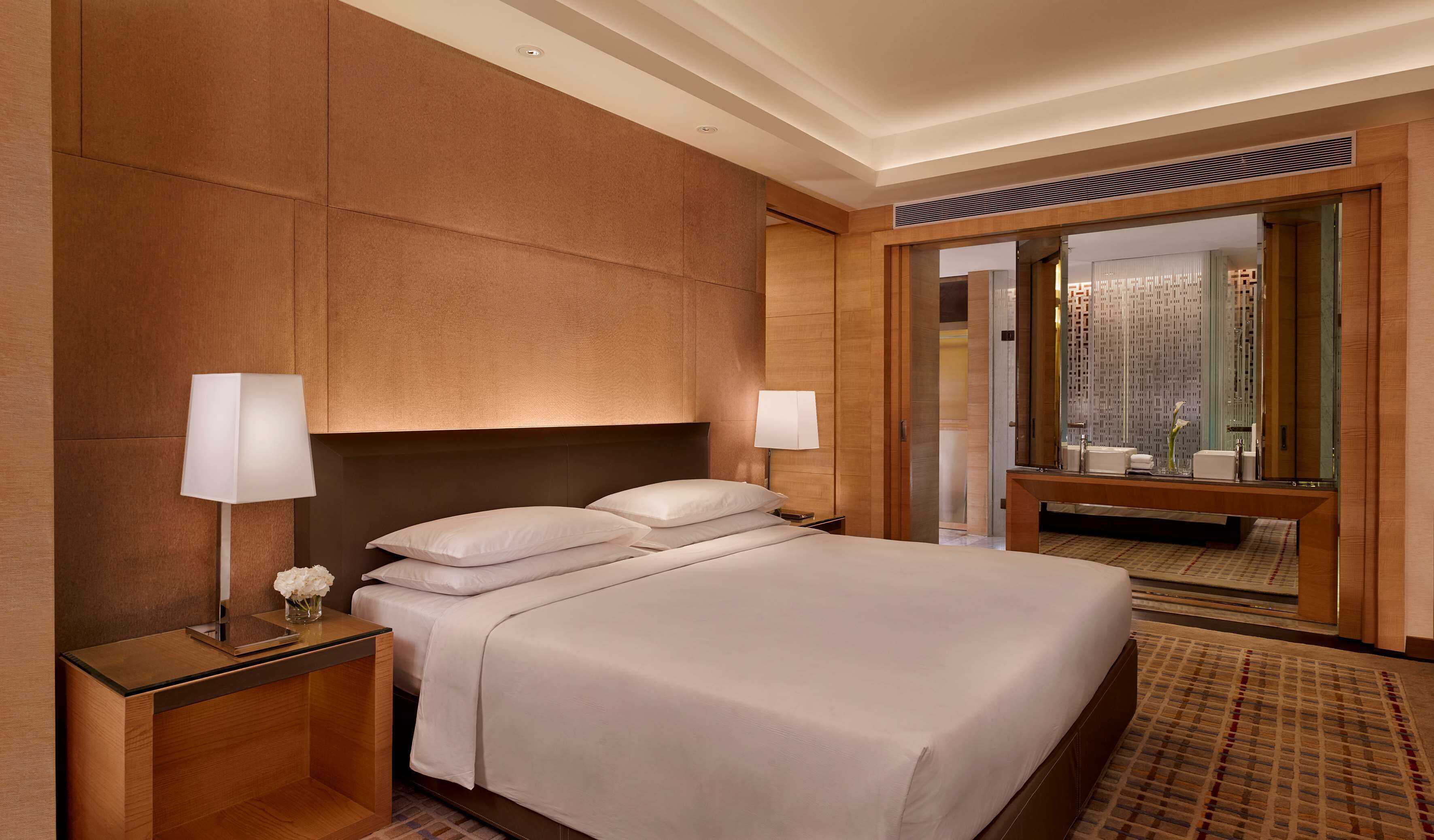 Une chambre d’hôtel avec un grand lit, deux lampes de chevet, une table de nuit ornée d’un vase de fleurs et un grand miroir qui reflète la salle de bain.