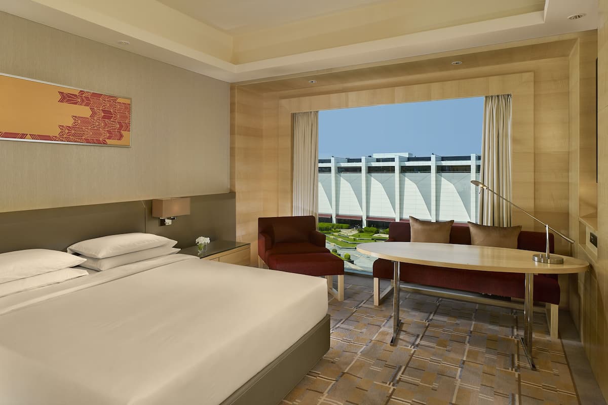 A hotel room with a large bed, a desk, a chair, and a view of a building through the window.