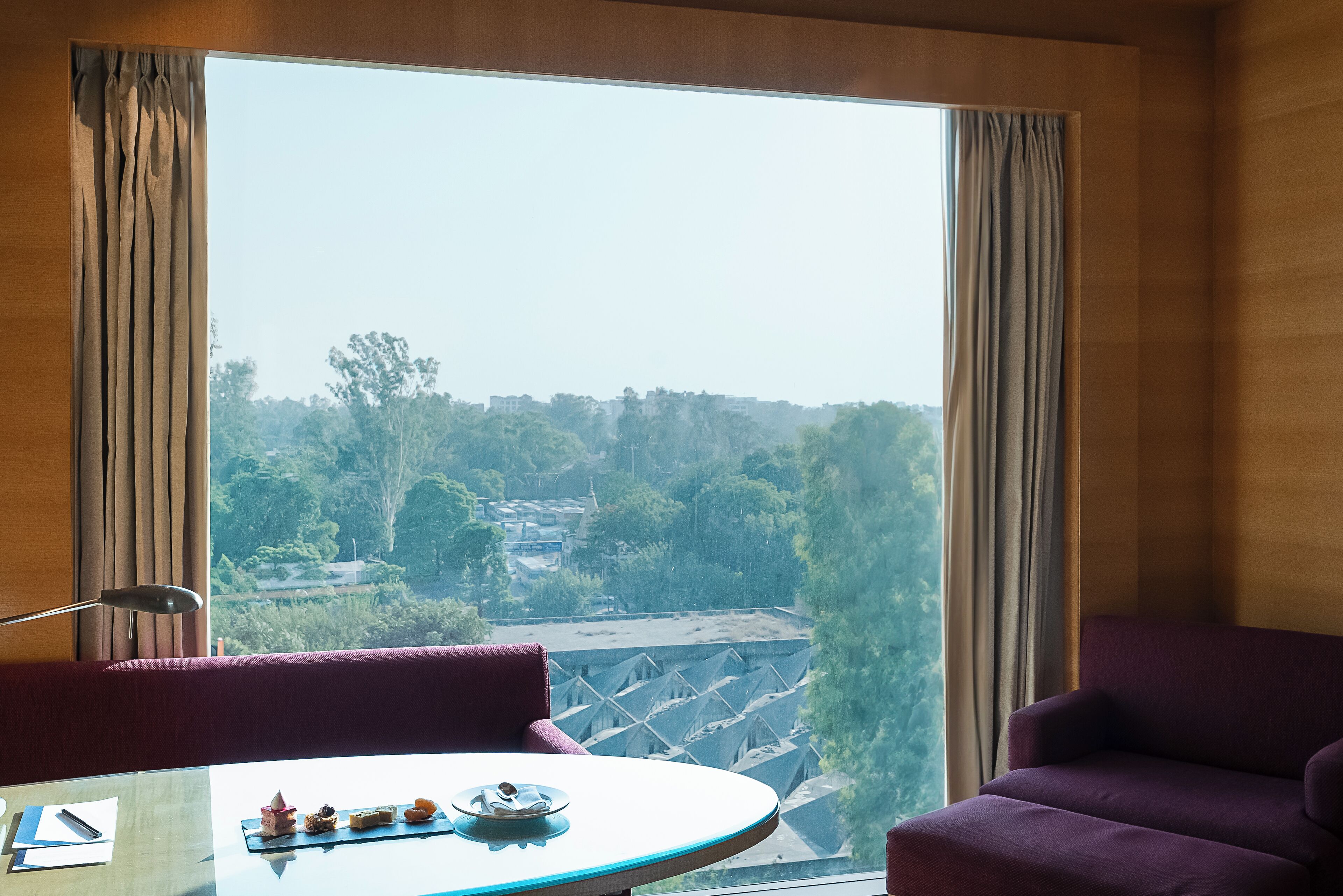 A room with a large window, a table with a plate of pastries, a chair, and a lamp.