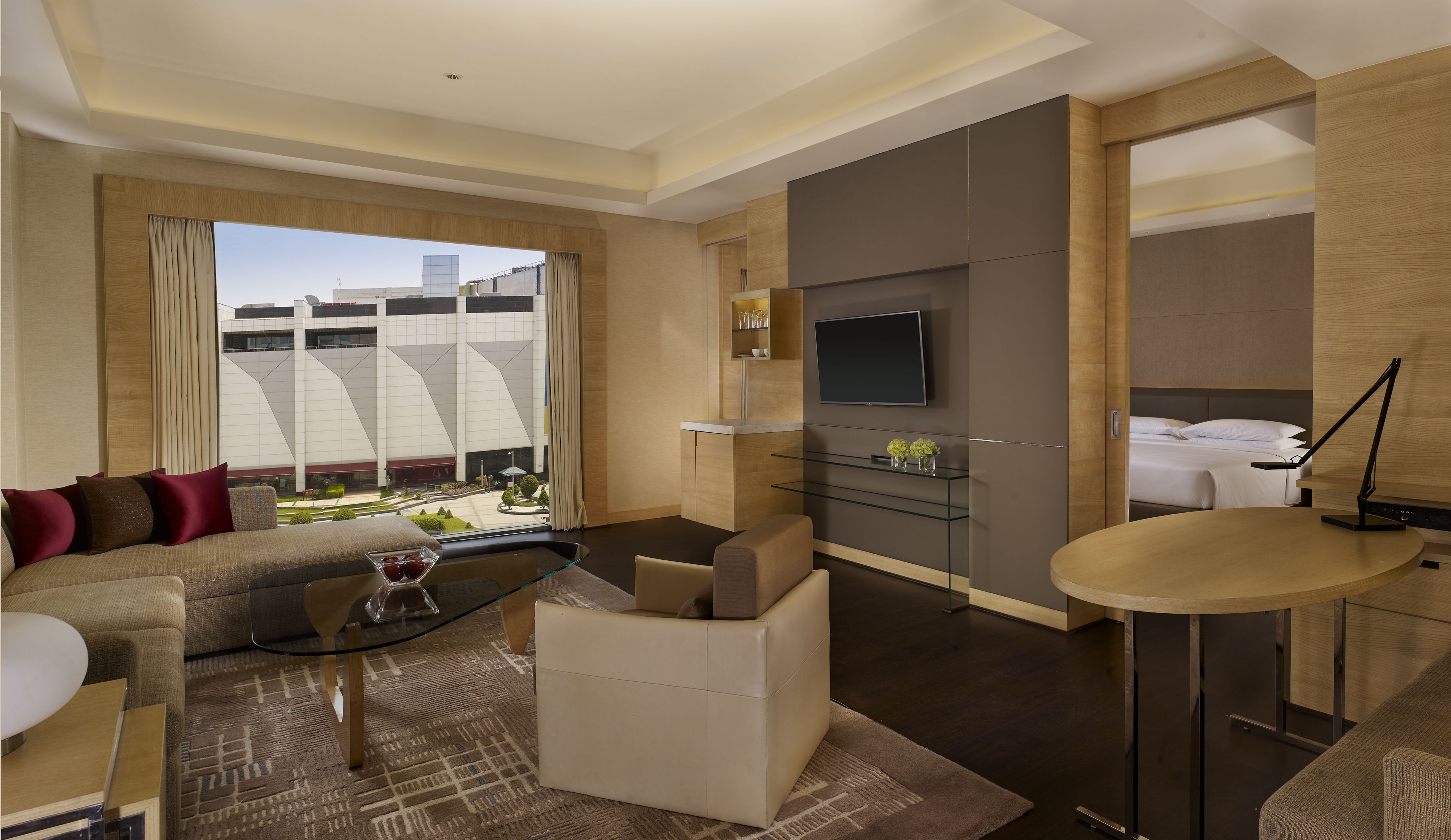 A modern hotel room with a sofa, a glass coffee table, a TV, a small dining table, and a view of the cityscape.