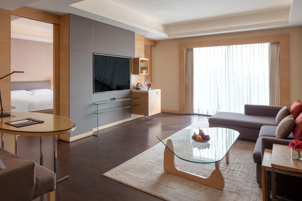 A modern hotel room with a flat-screen TV, a sofa, a glass coffee table, and a bed with a headboard.