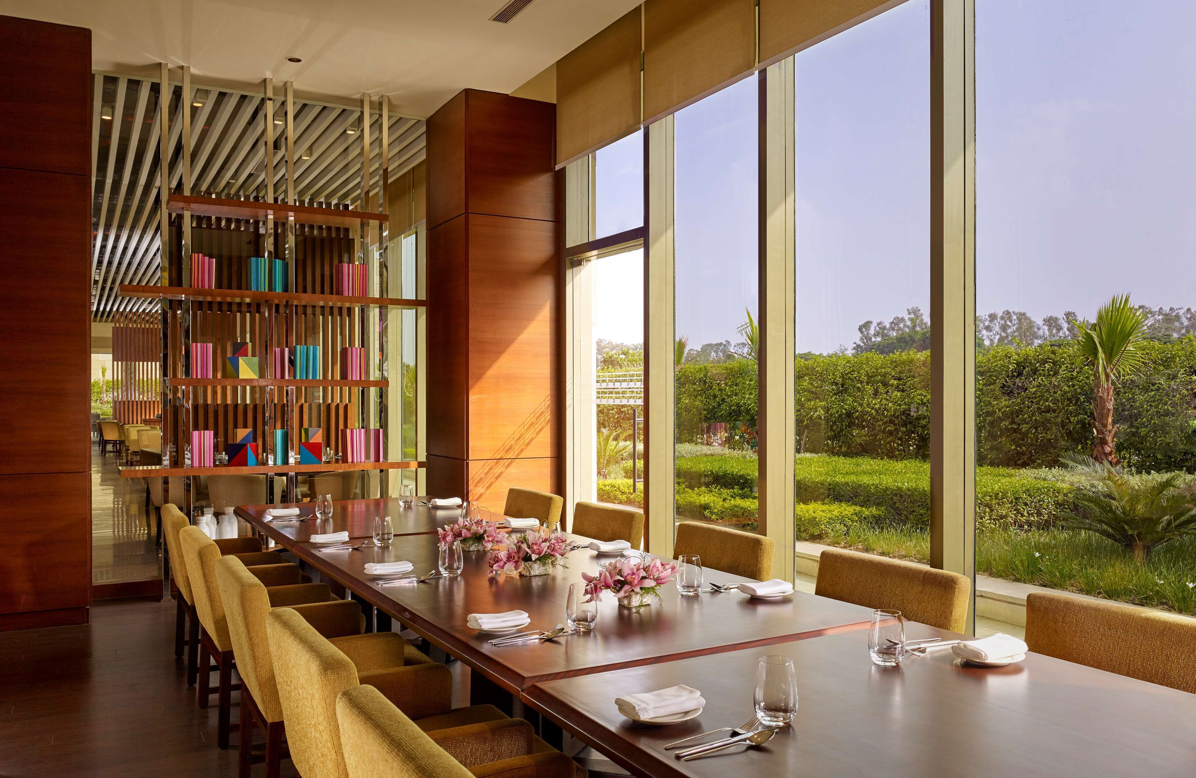 A dining area with a long table set for six, large windows offering a view of a well-manicured garden, and a modern bookshelf with colorful books.