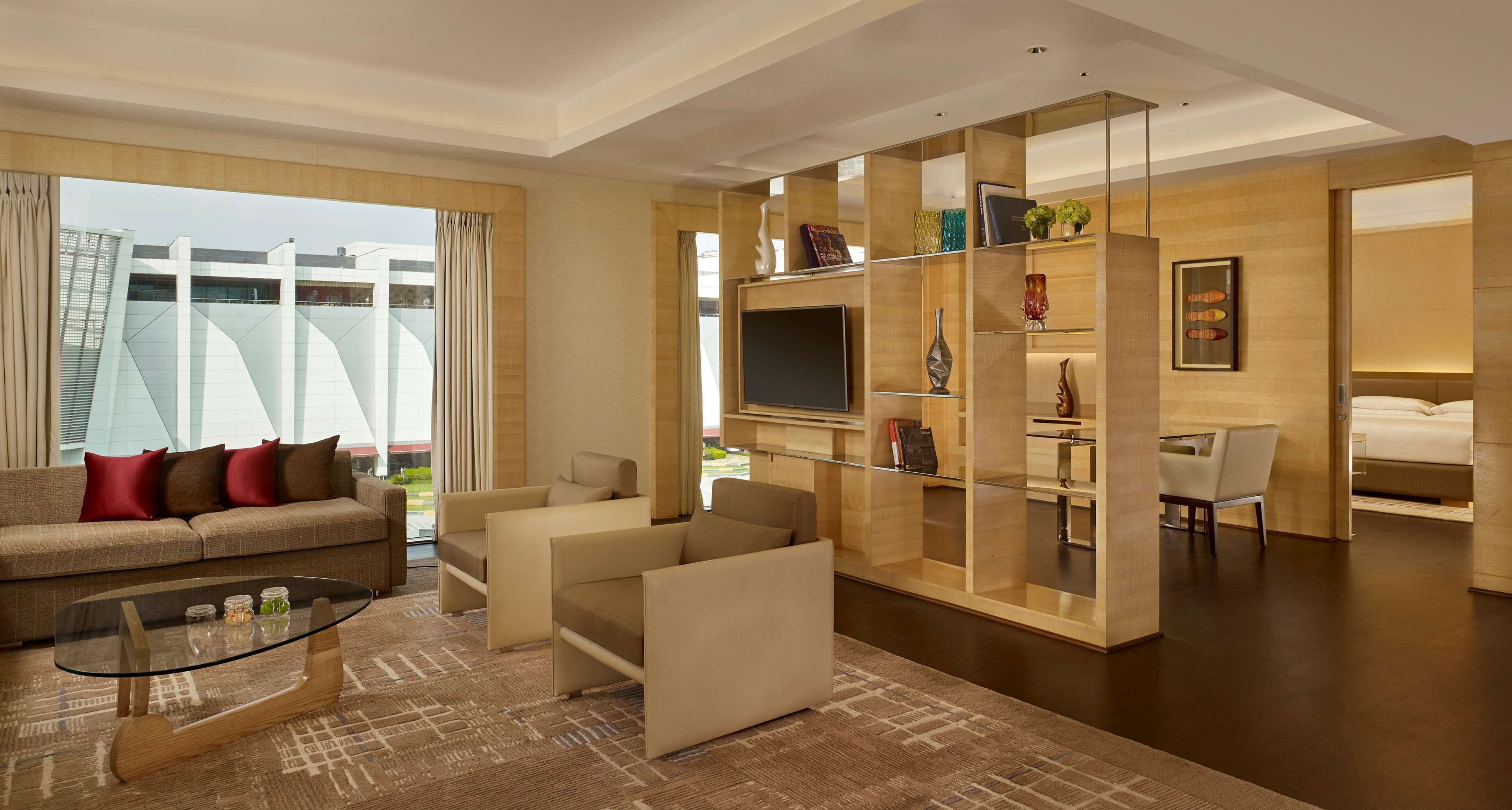A modern hotel room with a sofa, armchairs, a glass coffee table, a TV, and a bookshelf.
