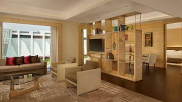 A modern hotel room with a sofa, armchairs, a glass coffee table, a TV, and a bookshelf.