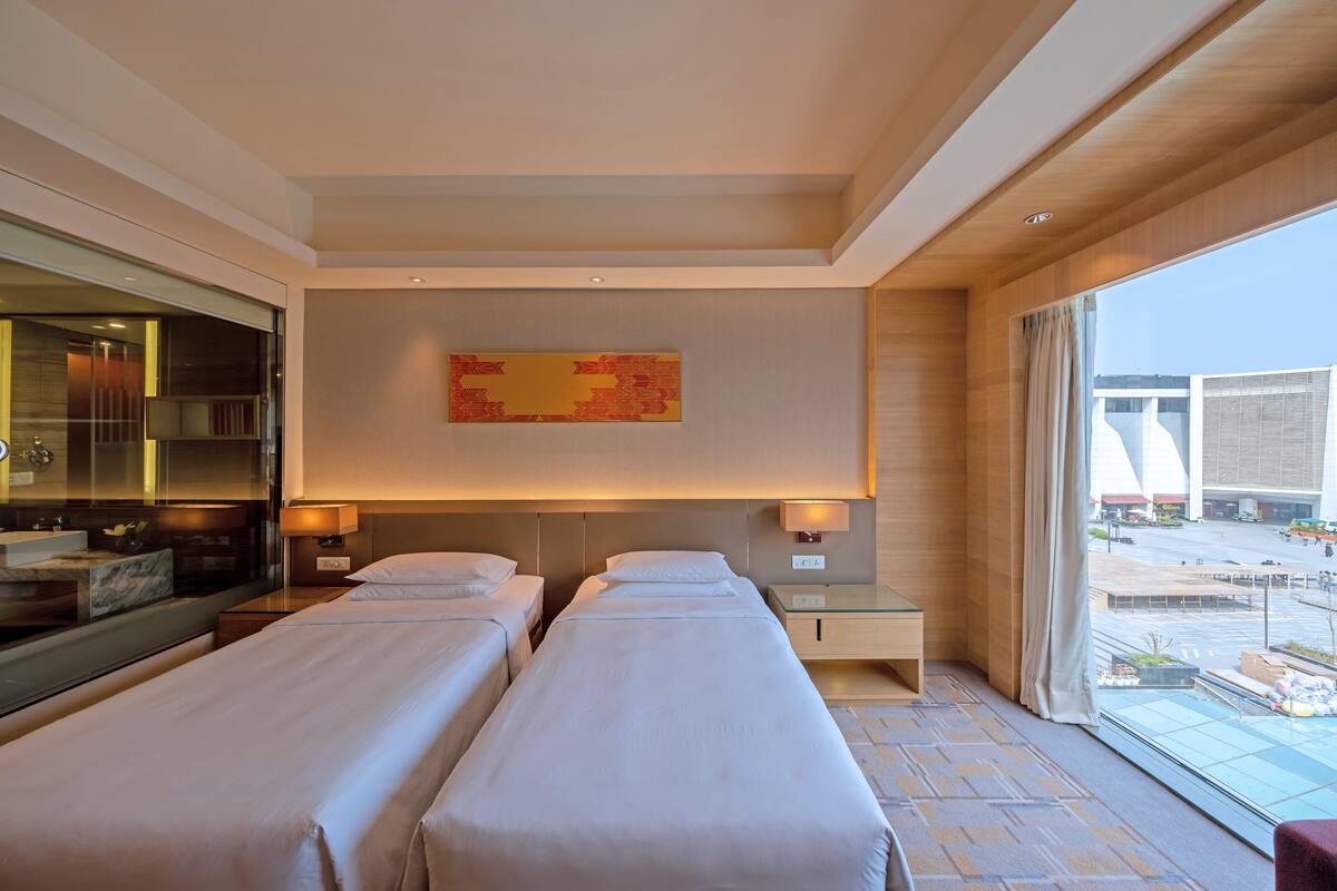 A hotel room with two beds, a large window, a bathroom with a mirror, and a wall-mounted artwork.