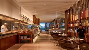 Interior moderno de un restaurante con una larga barra, chefs preparando comida y una zona de bar con estanterías llenas de botellas.