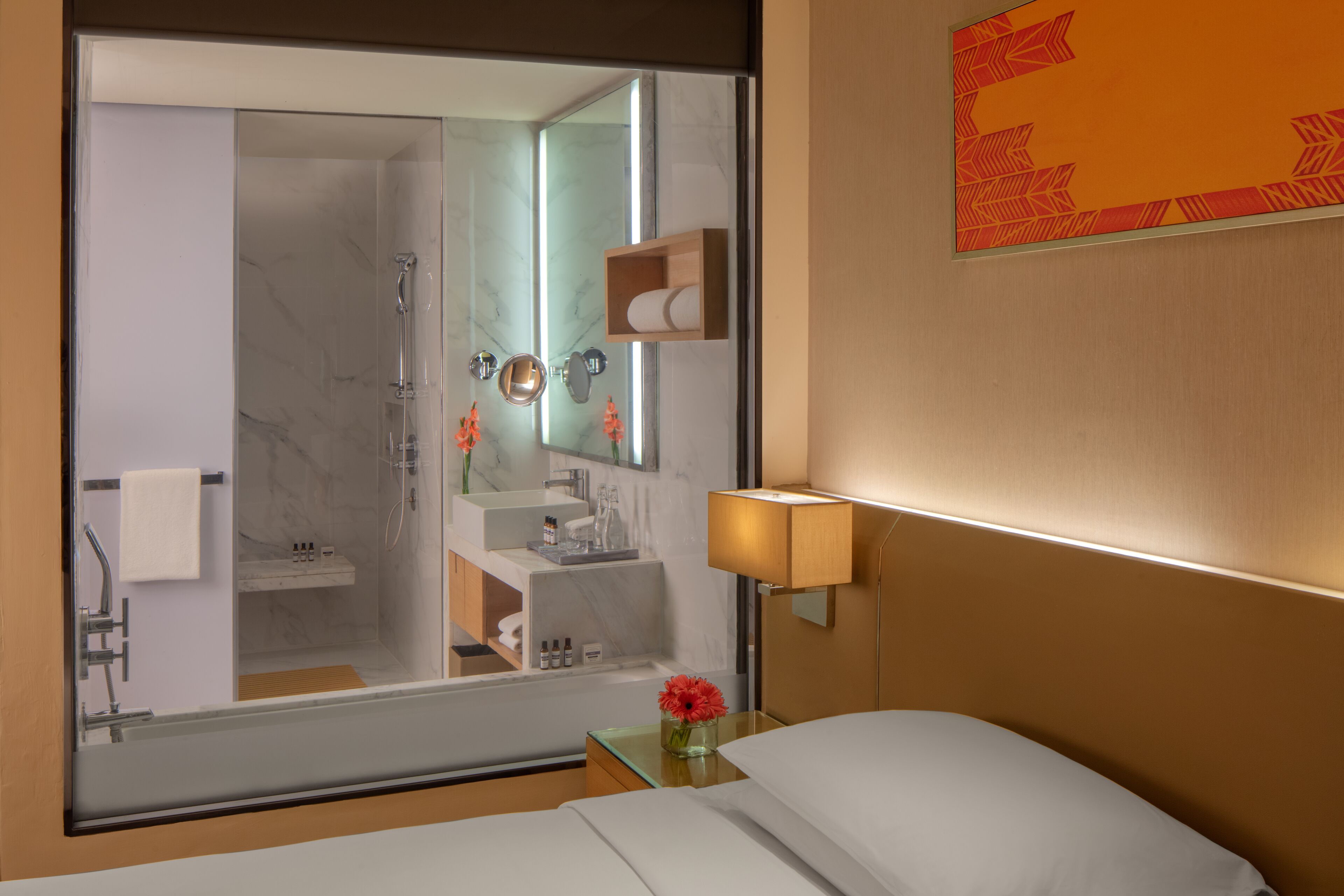 A hotel room with a large mirror reflecting the bathroom, a bedside table with a lamp, a bed, and a small vase with flowers.