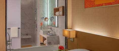 A hotel room with a large mirror reflecting the bathroom, a bedside table with a lamp, a bed, and a small vase with flowers.