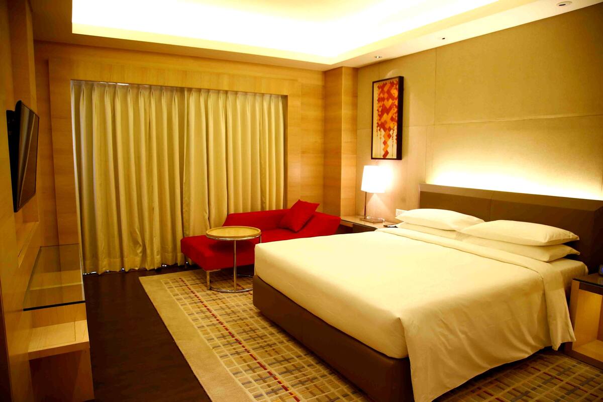A hotel room with a large bed, a red chair, a small round table, a lamp, and a painting on the wall.