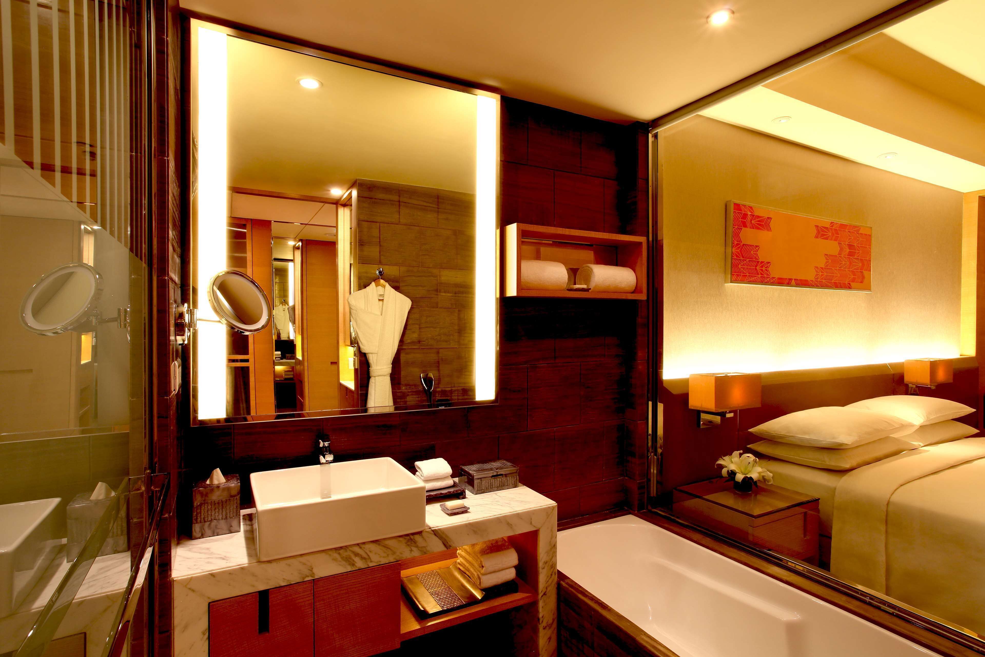 A modern bathroom with a large mirror, a sink, a bathtub, and a bedside table with a lamp.