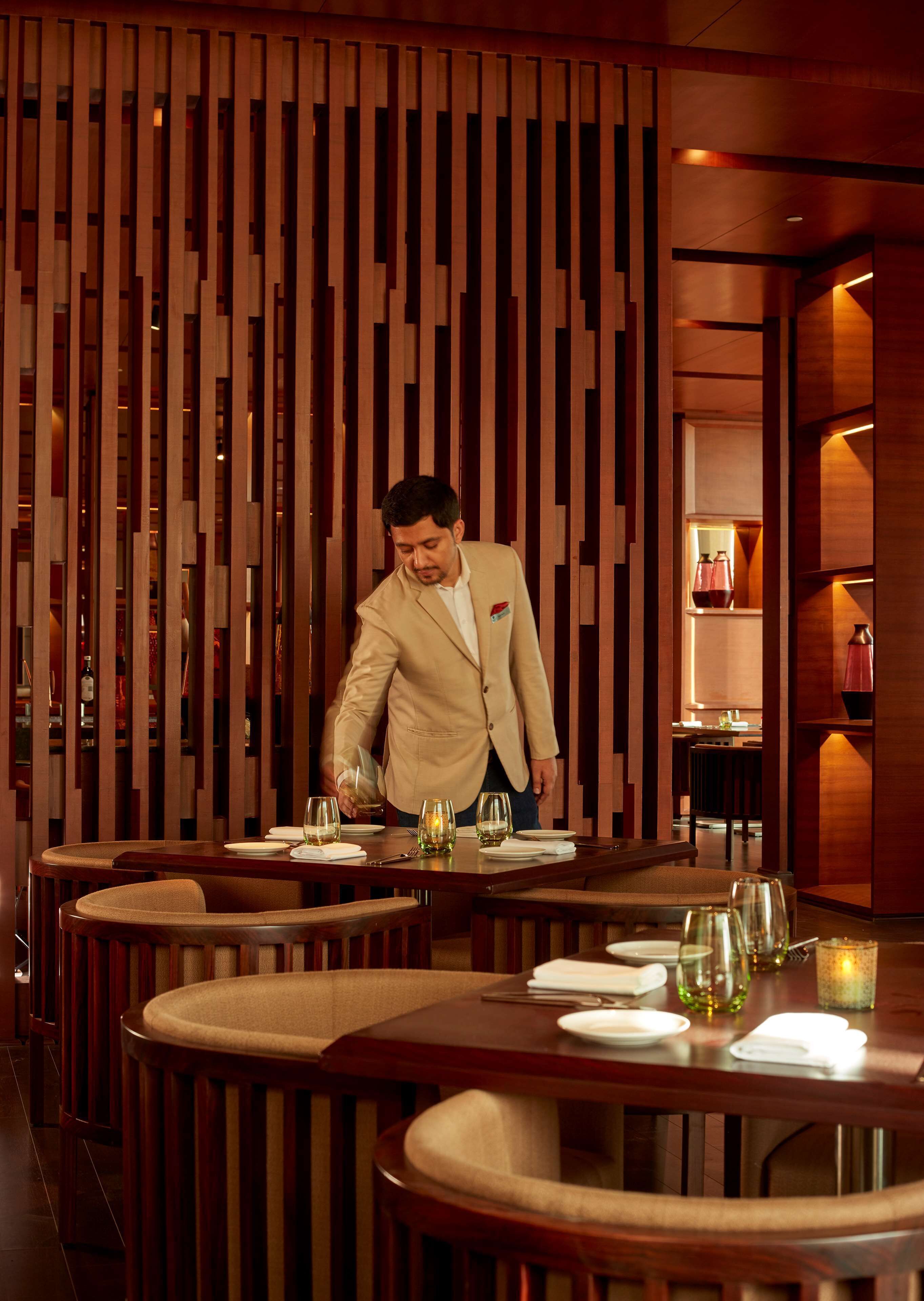A man in a beige suit serving at a restaurant with wooden paneling and round tables.