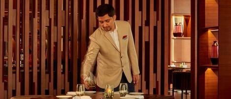 A man in a beige suit serving at a restaurant with wooden paneling and round tables.