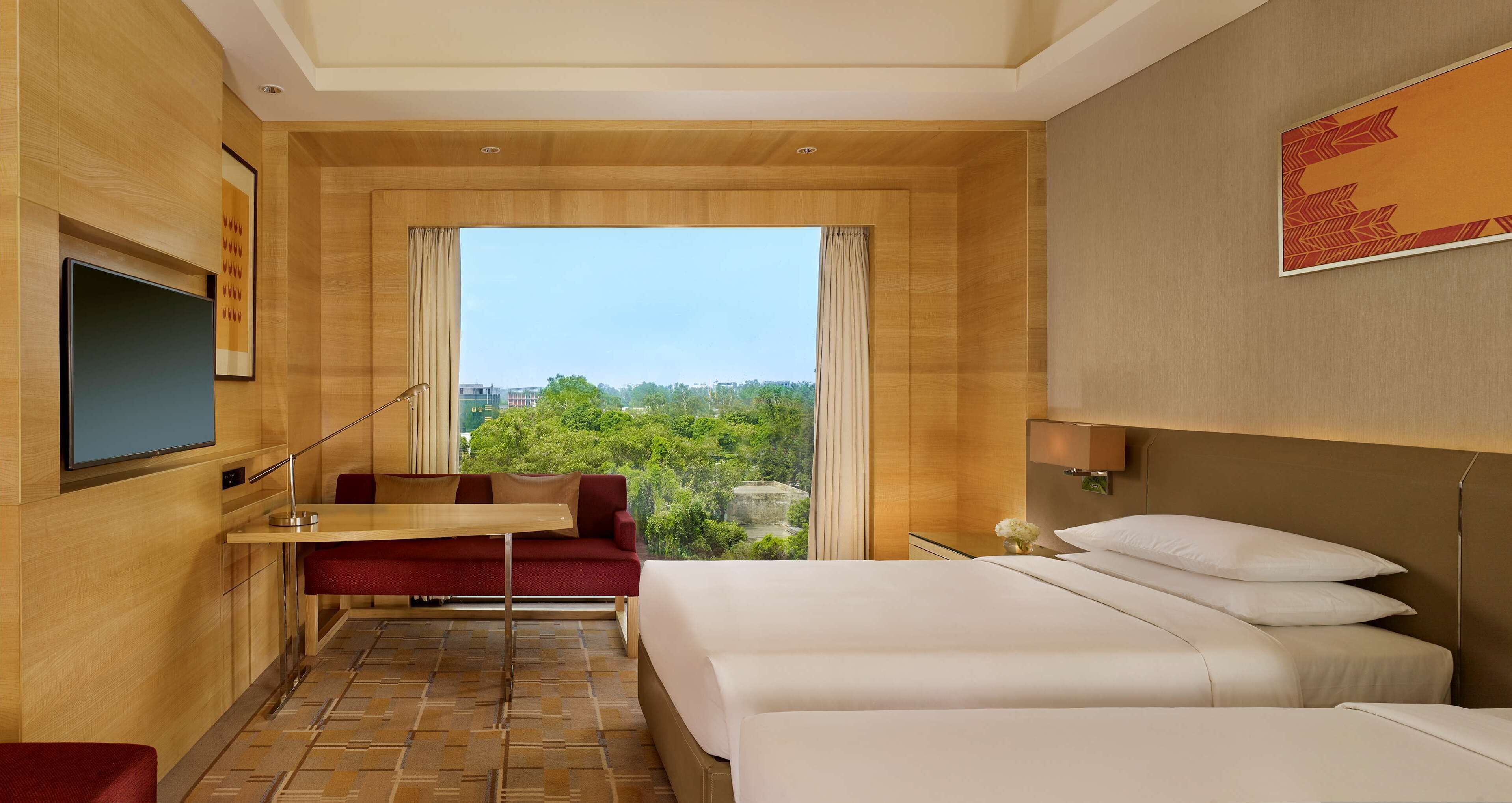 A hotel room with a bed, a desk, a chair, a TV, and a window with a view of greenery.