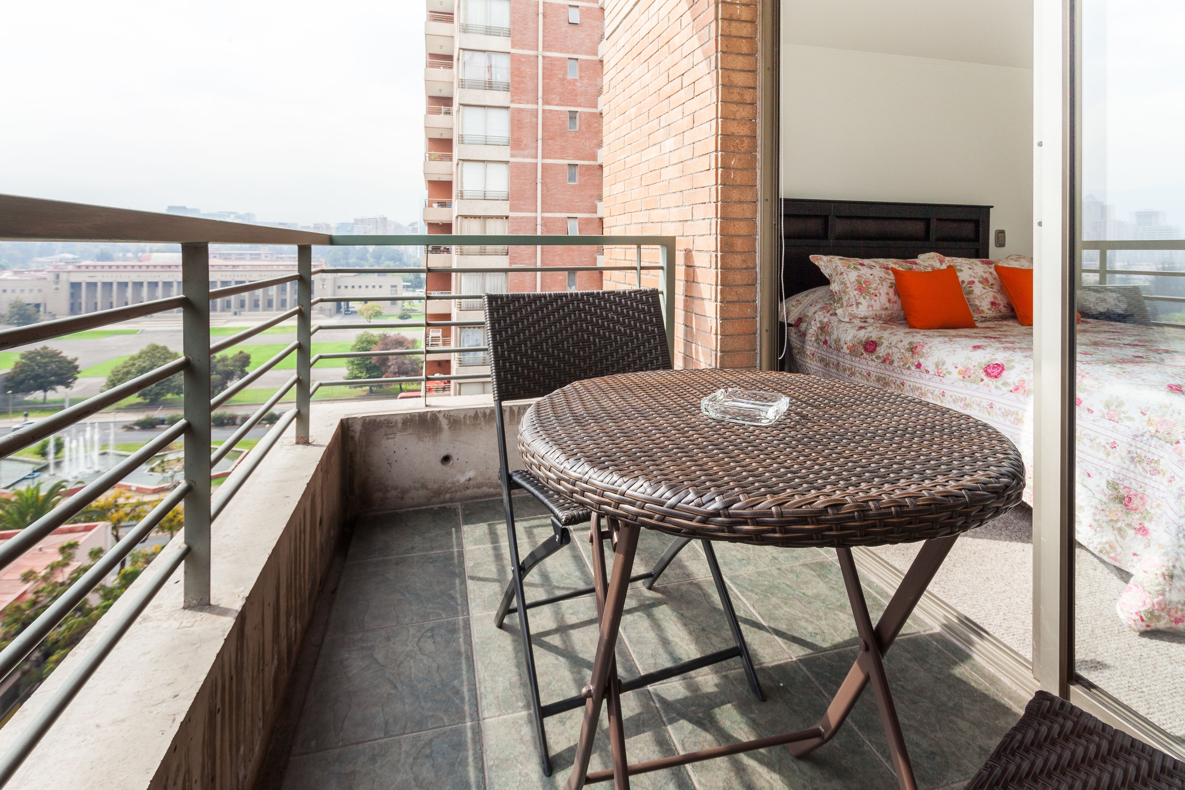 Een balkon met een rieten tafeltje en stoel, een bed met bloemenbedekking en uitzicht op een stadsgezicht.