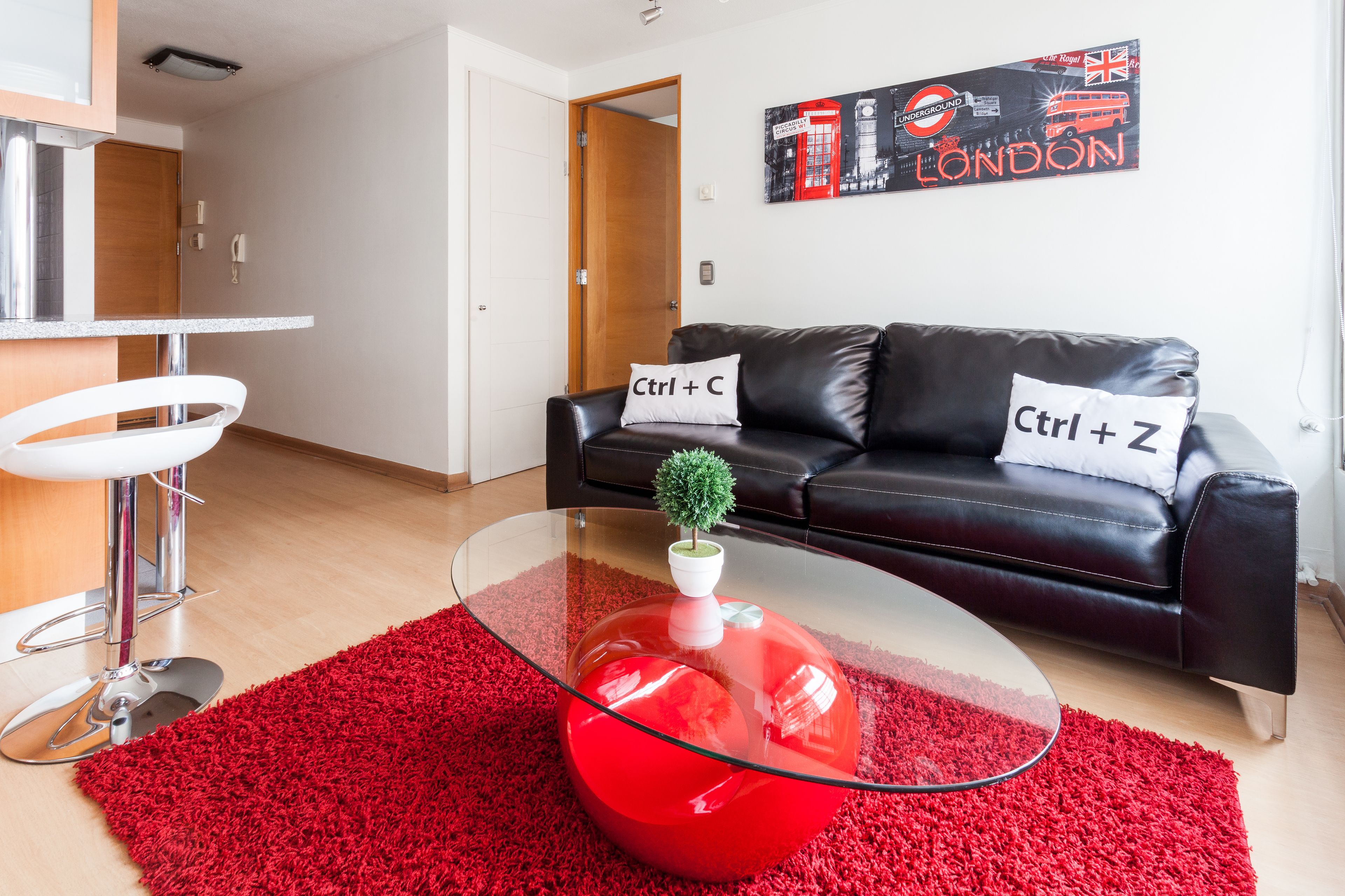 Een moderne woonkamer met een glazen salontafel, een rood tapijt en een zwarte leren bank met decoratieve kussens.
