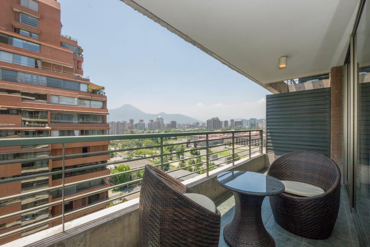 Un balcón con sillas de mimbre y una mesa, con vistas a un paisaje urbano con edificios y una montaña.