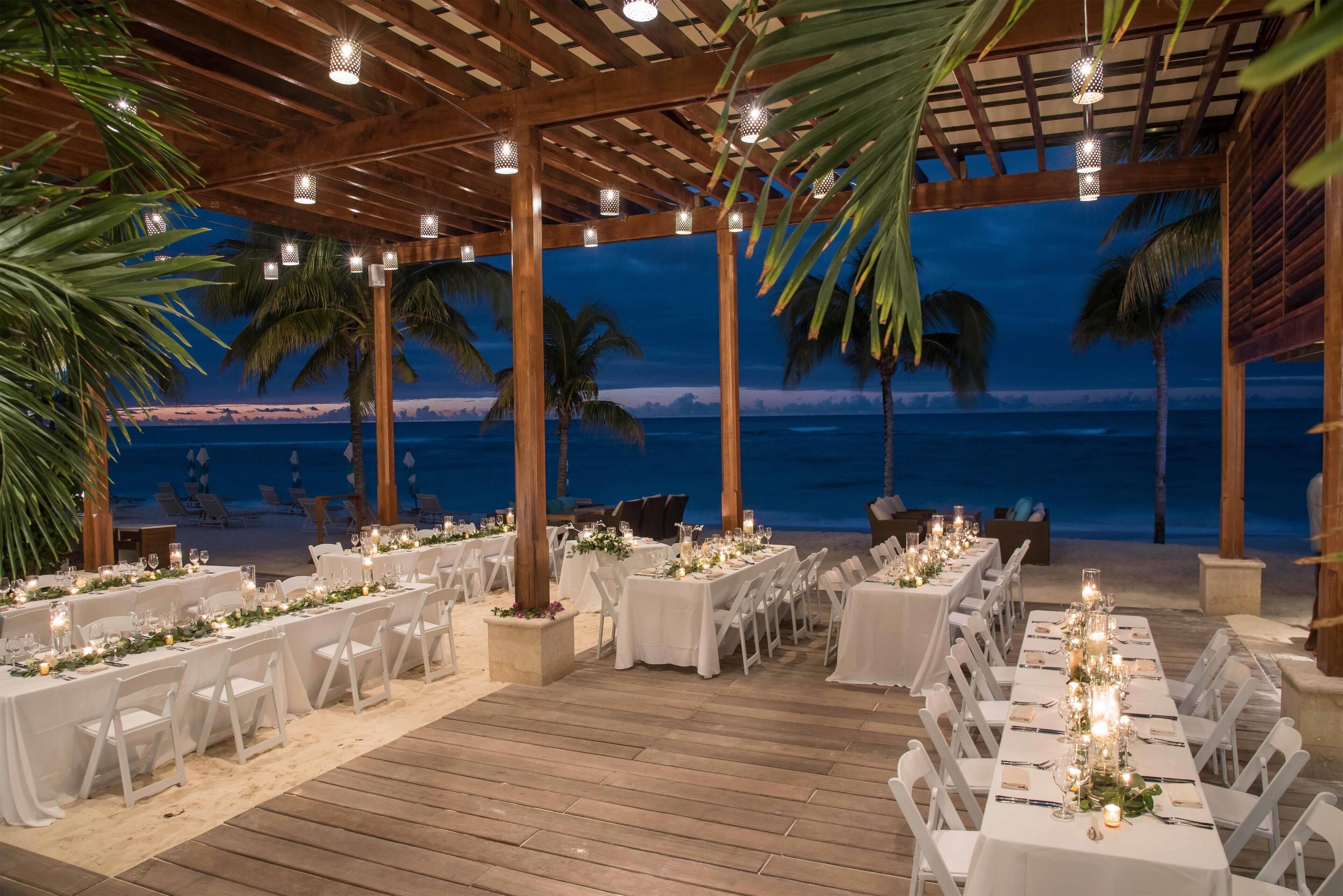An outdoor dining area with tables set for a wedding, overlooking the beach at dusk.