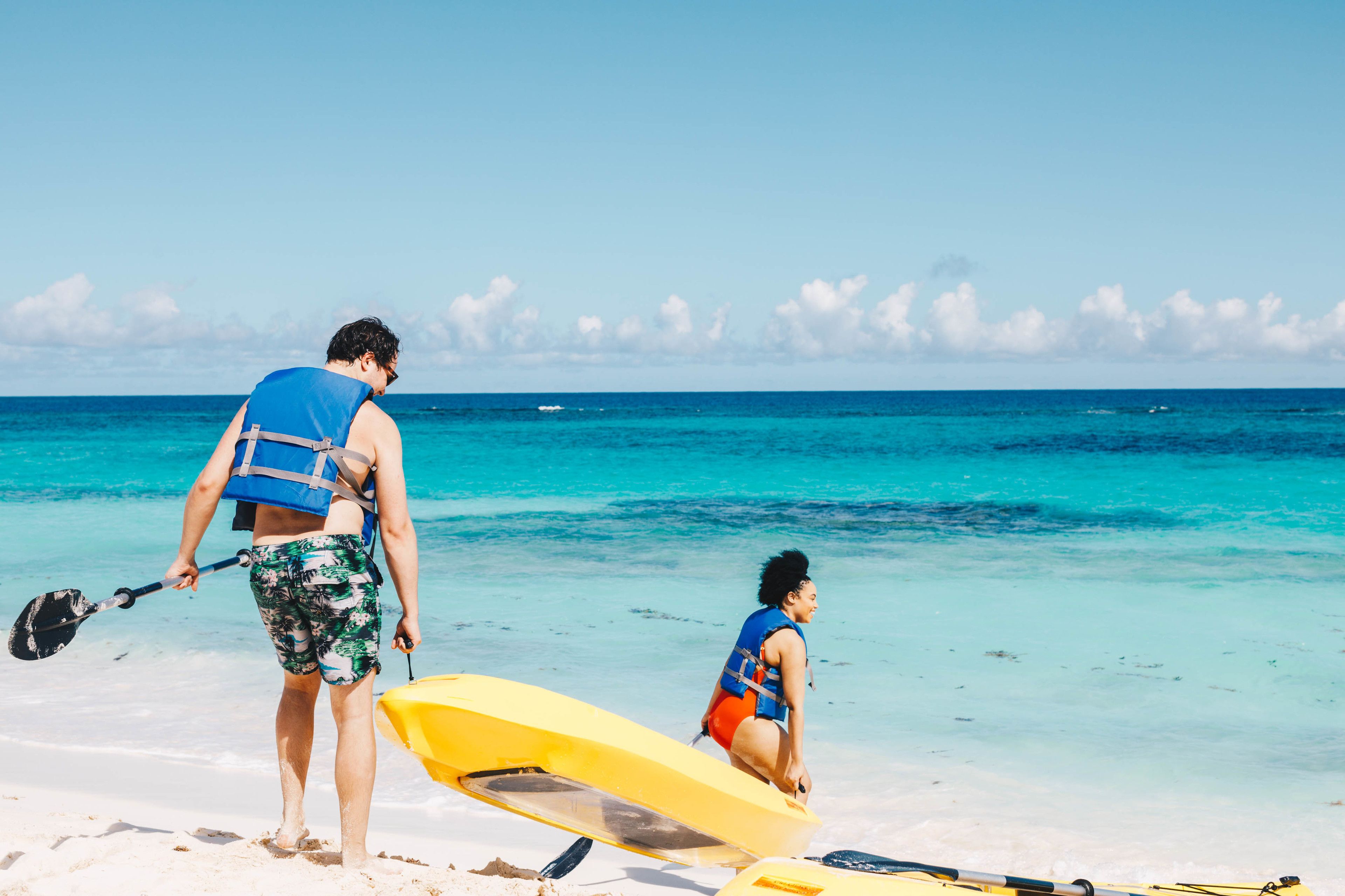 A beach scene with a person holding a paddle and a yellow kayak, and another person in a blue life jacket and red shorts.