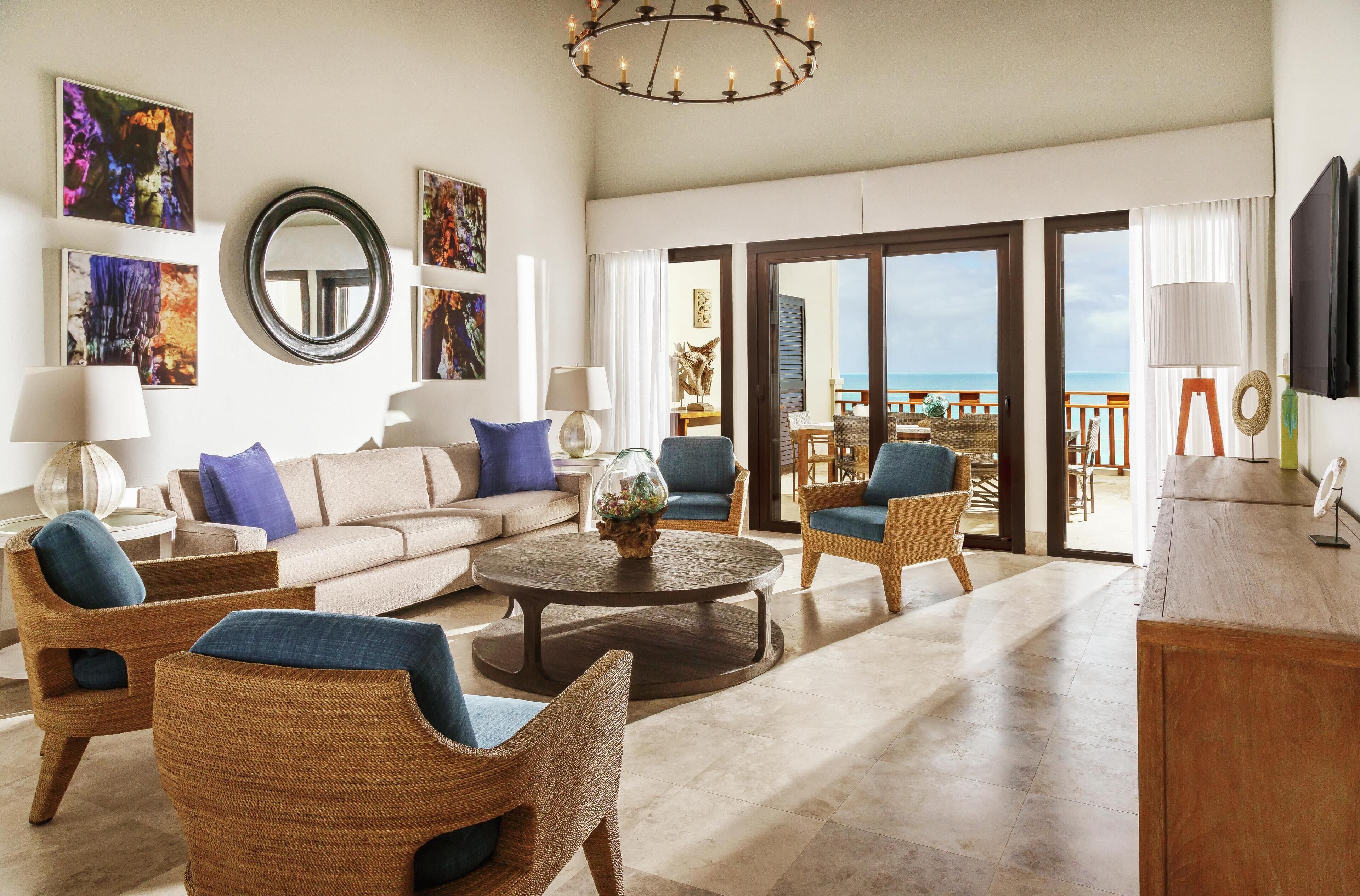 A spacious living room with a large sofa, two armchairs, a coffee table, and a TV. There are framed pictures on the wall, a mirror, and a view of the ocean through the sliding glass doors.
