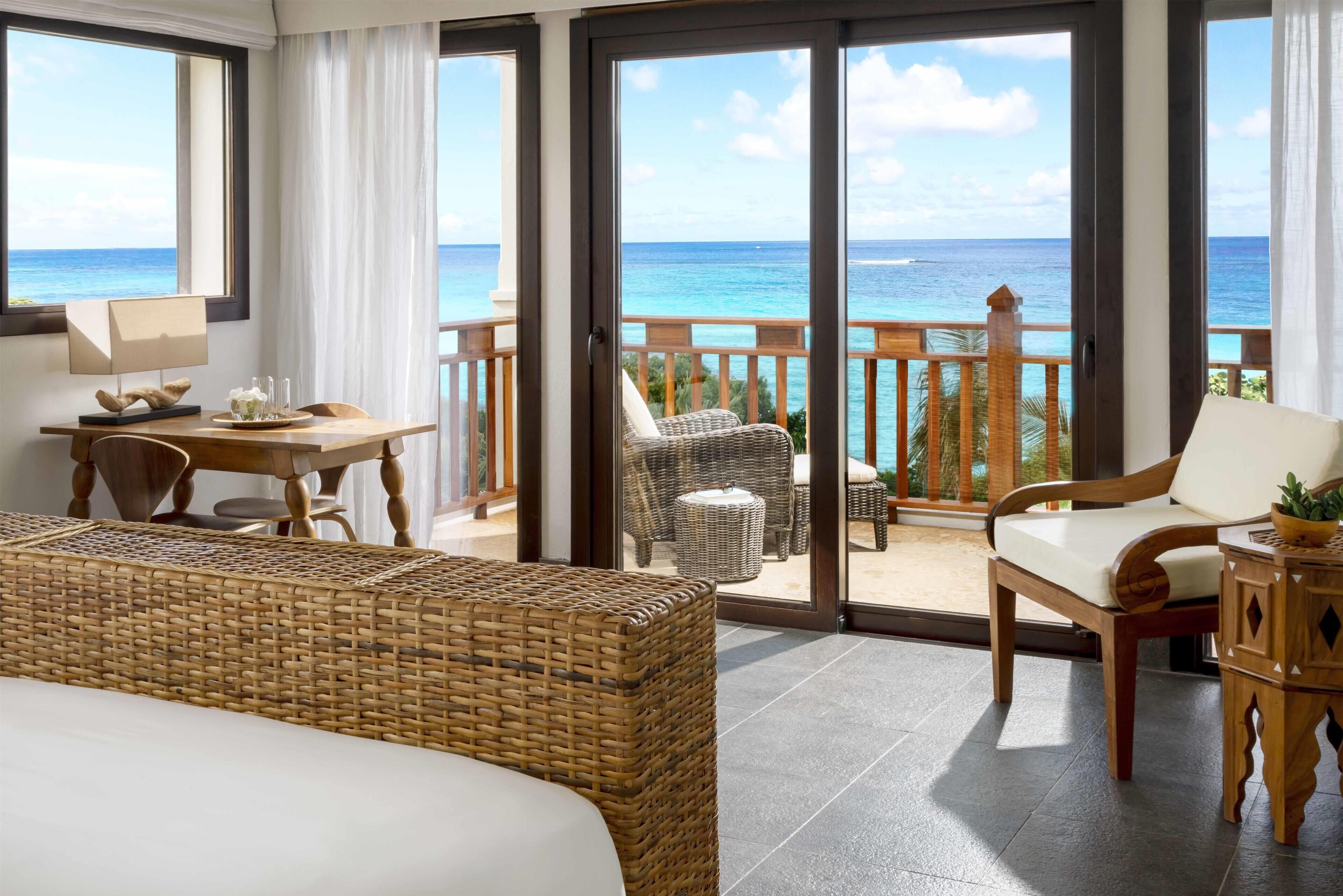 A room with a bed, a dining table with chairs, a wicker sofa, and a balcony with a view of the ocean.