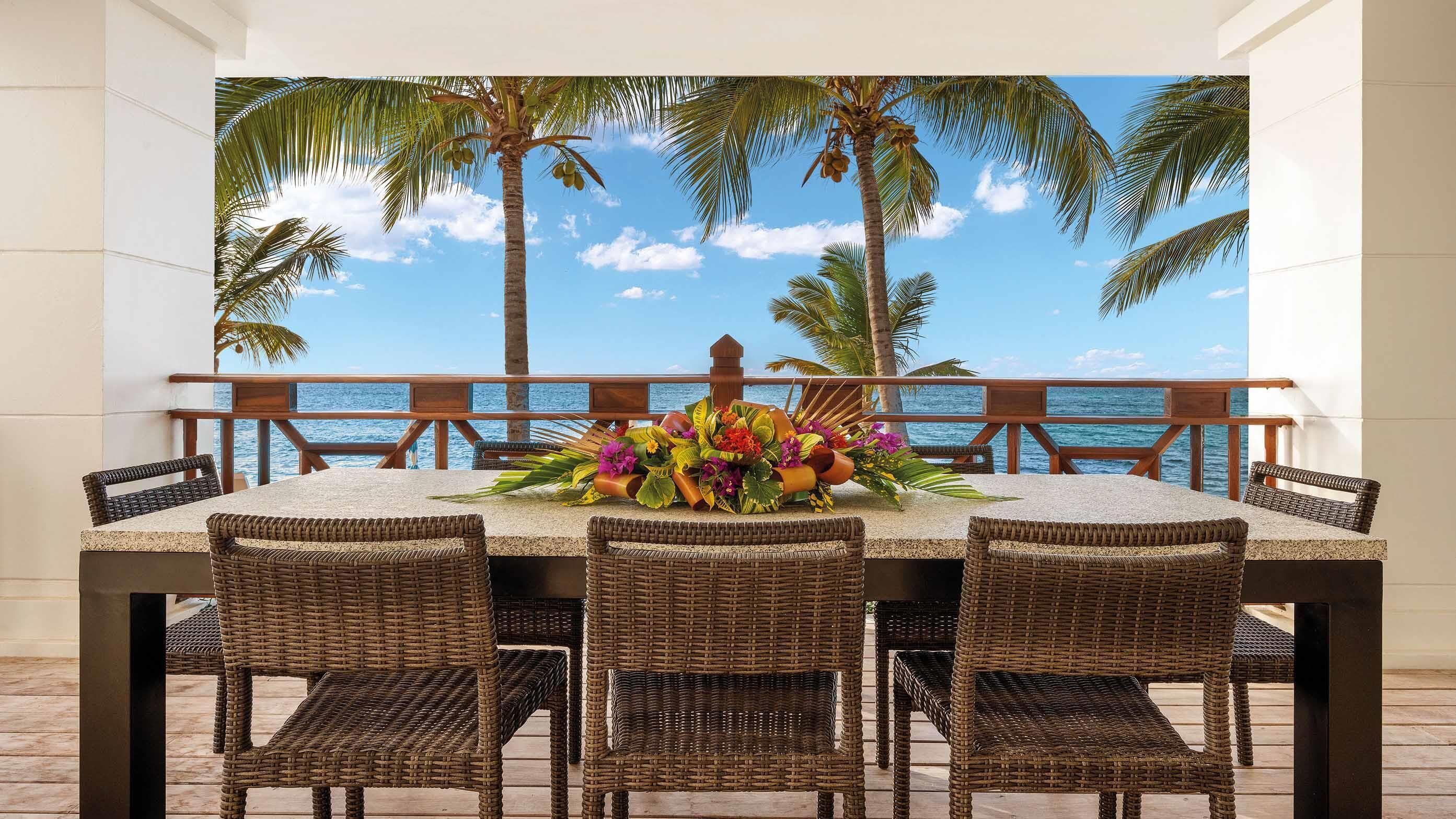 Un coin repas avec une table dressée pour quatre personnes, entouré de chaises en osier, et offrant une vue sur l’océan et les palmiers.