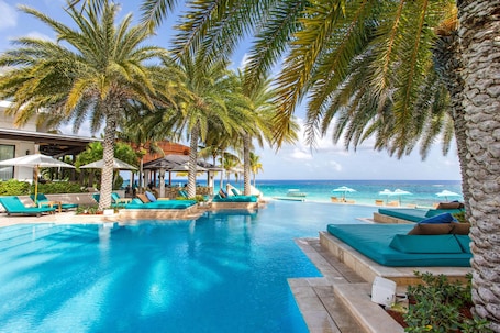 A pool area with lounge chairs and umbrellas, surrounded by palm trees and a view of the ocean.
