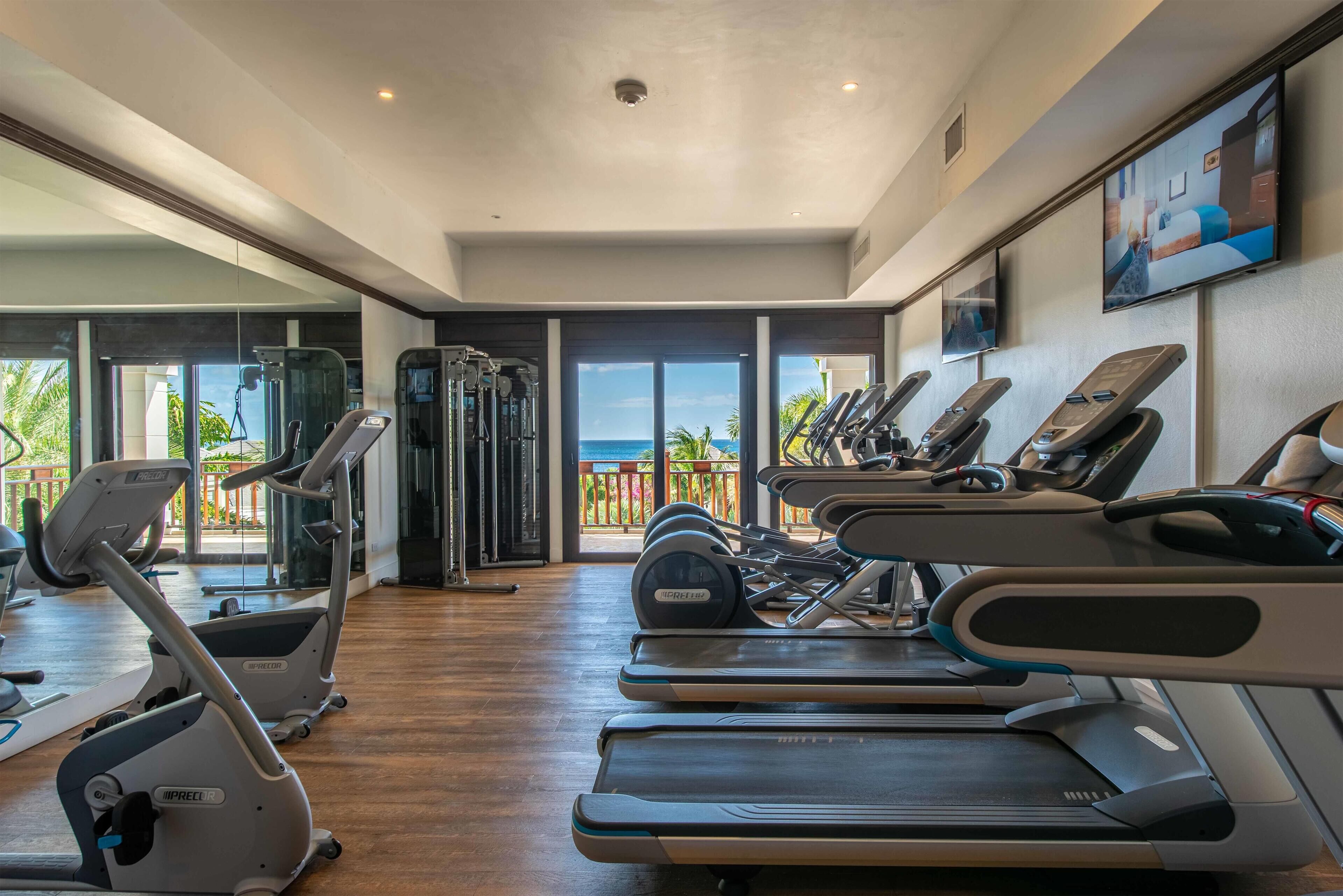 A gym with treadmills, elliptical machines, and free weights, featuring large windows and a view of the ocean.