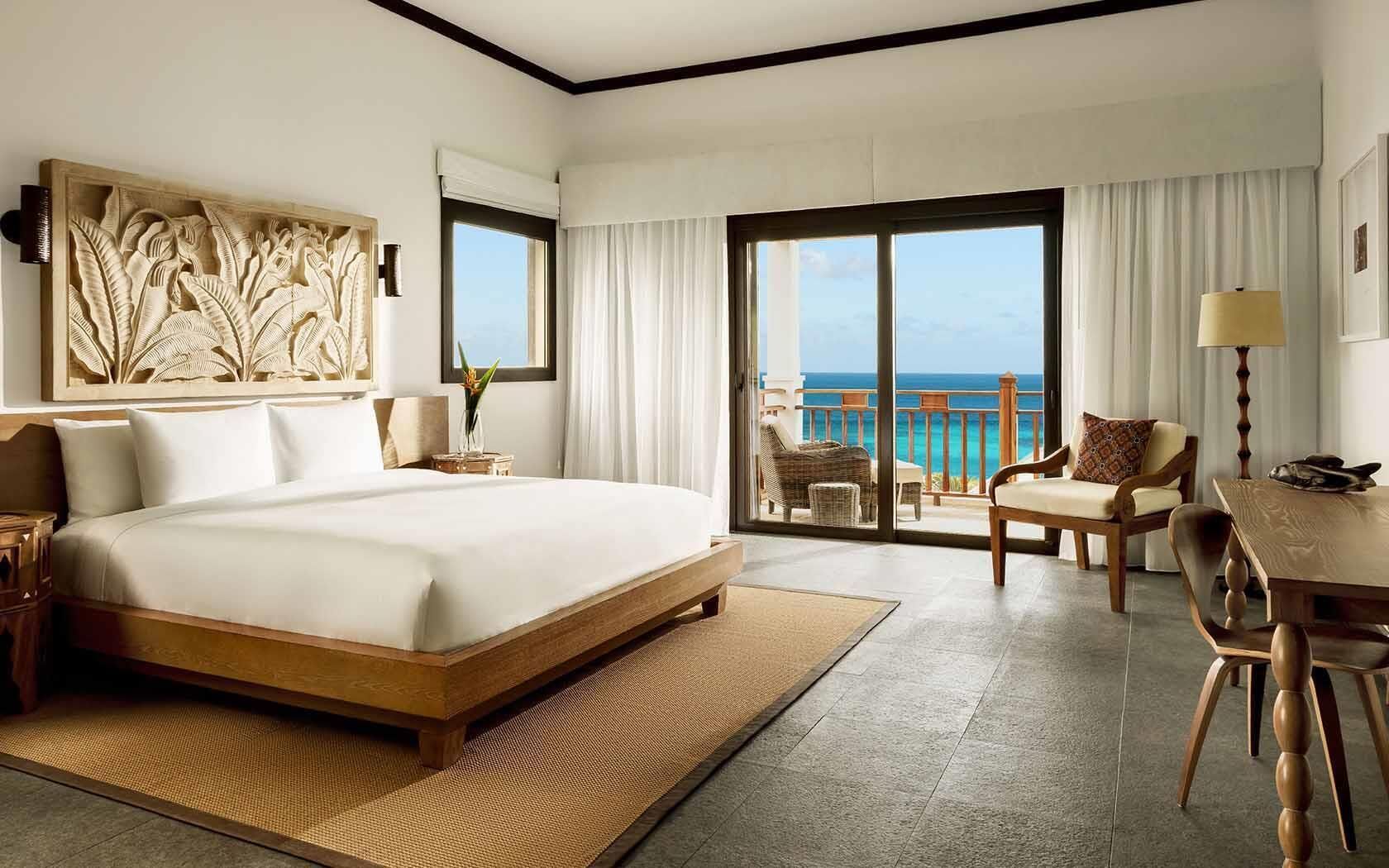 A spacious hotel room with a large bed, a balcony with a view of the ocean, and a dining area with wooden furniture.