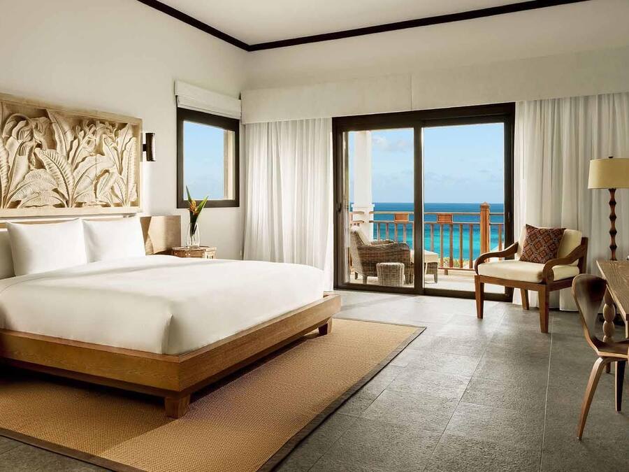 A spacious hotel room with a large bed, a balcony with a view of the ocean, and a dining area with wooden furniture.