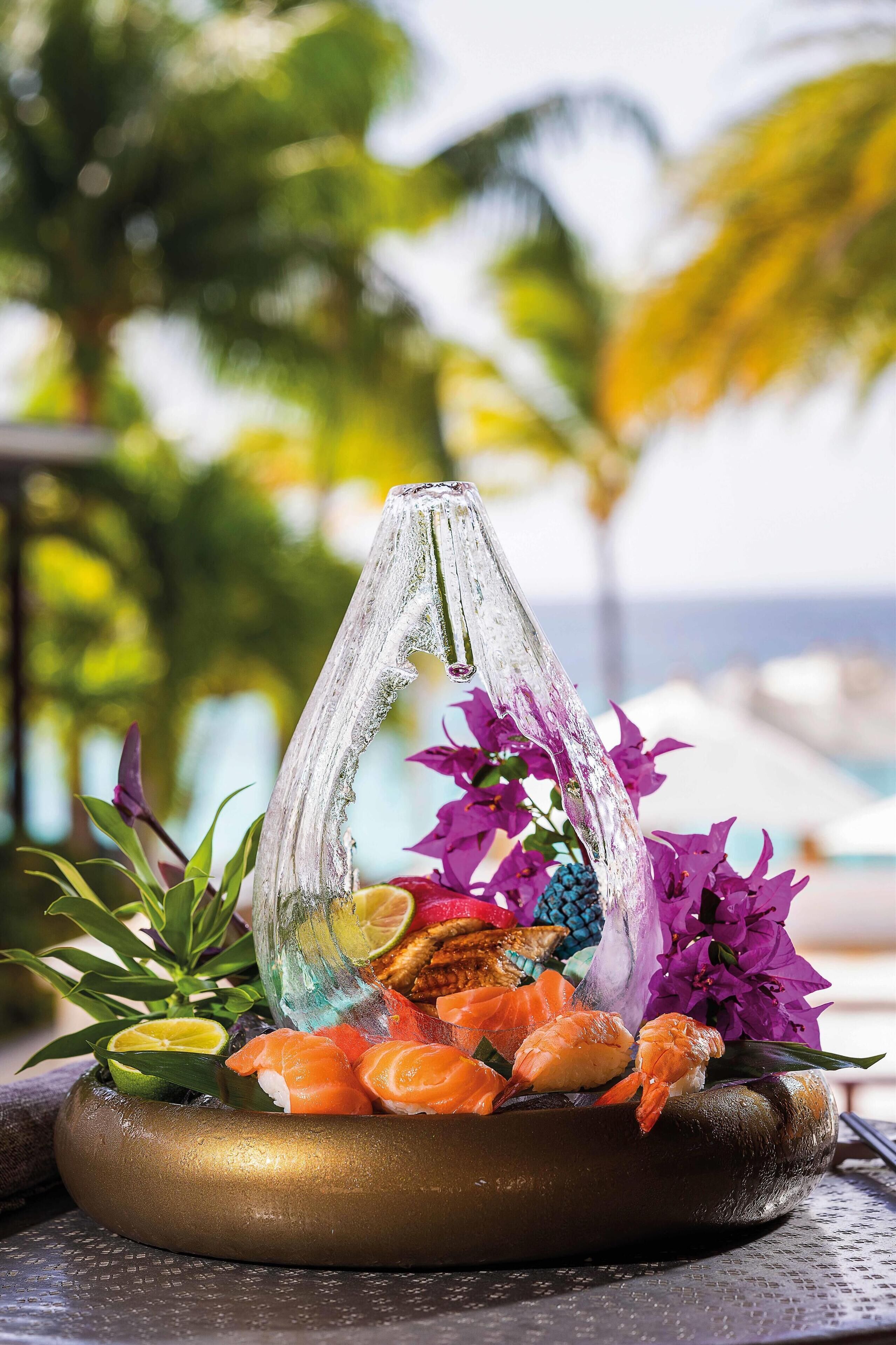 A sushi platter with shrimp and other seafood, garnished with purple flowers and a glass vase, set on a round, dark-colored tray.