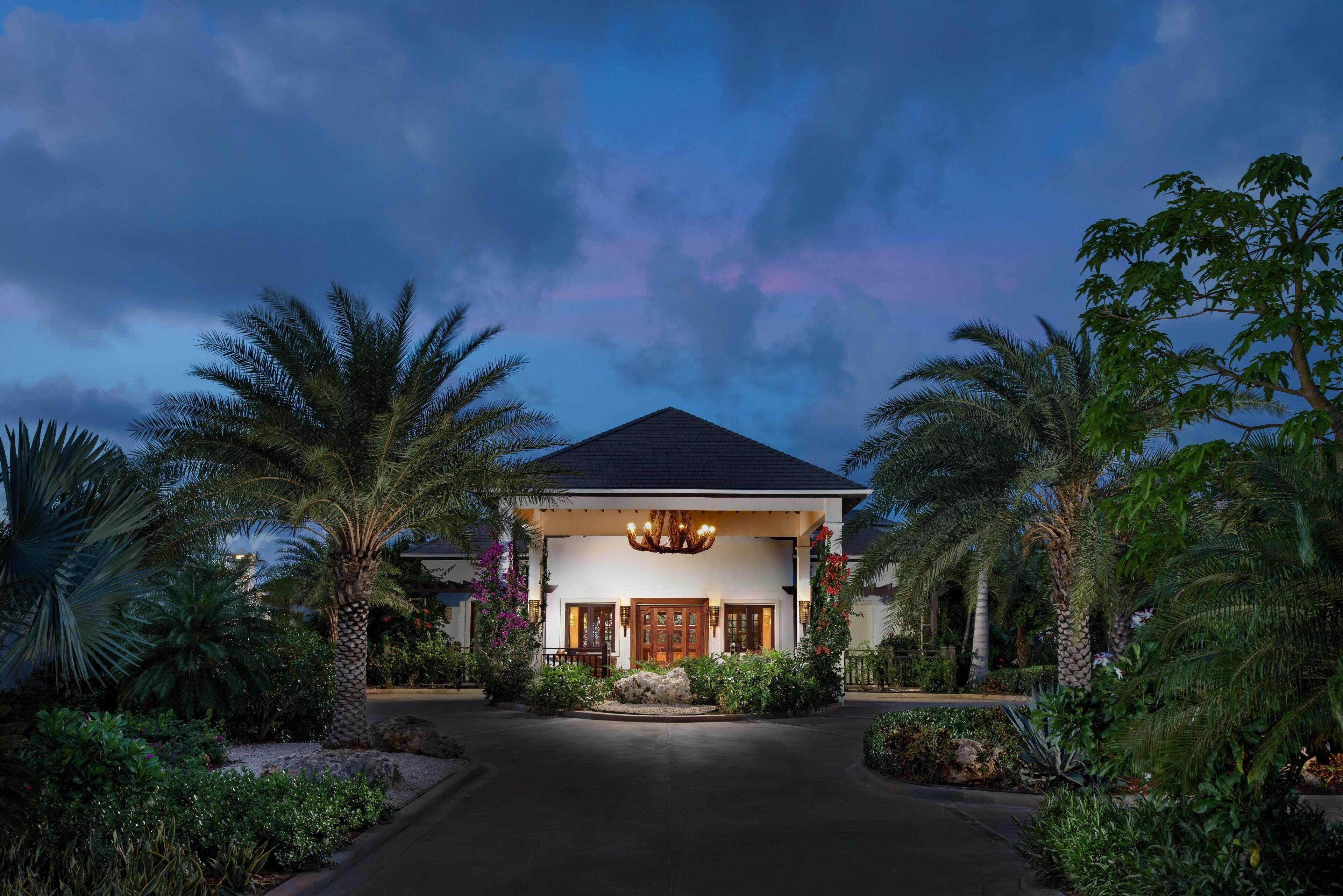 A two-story house with a dark roof, a central entrance with a chandelier, and a garden with palm trees and shrubs.