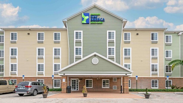 Ein mehrstöckiges Hotel mit grüner Fassade, einem Schild für Extended Stay America Select Suites und einem Parkplatz mit mehreren Autos.