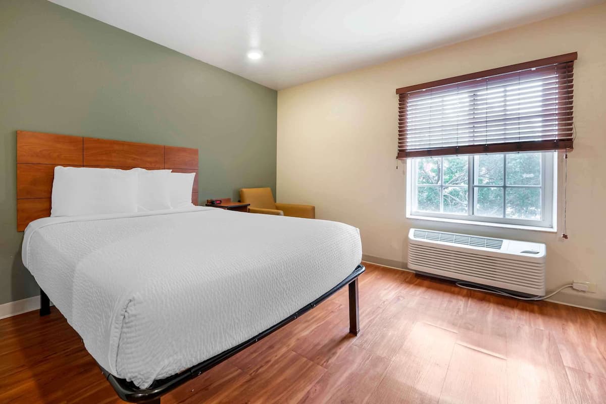 A bedroom with a bed, a chair, a window with blinds, and a wall-mounted air conditioner.