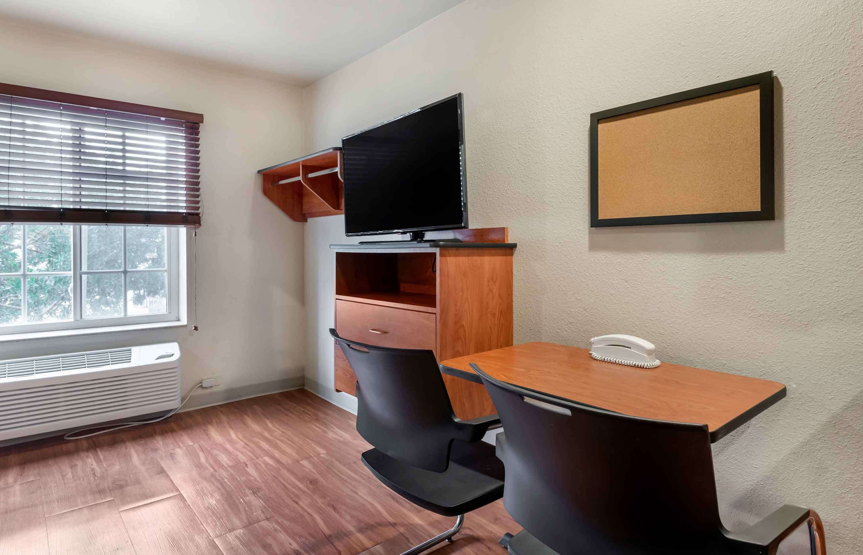 A small office space with a desk, two chairs, a television on a stand, a corkboard on the wall, and a window with blinds.