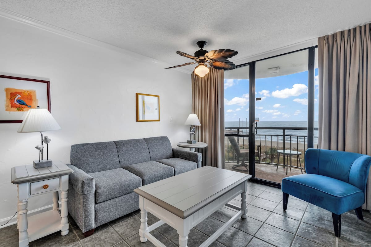 Habitación de hotel moderna con sofá gris, sillón azul, mesita de café blanca y ventilador de techo. Hay dos lámparas sobre mesitas auxiliares y un ventanal con vistas al océano.