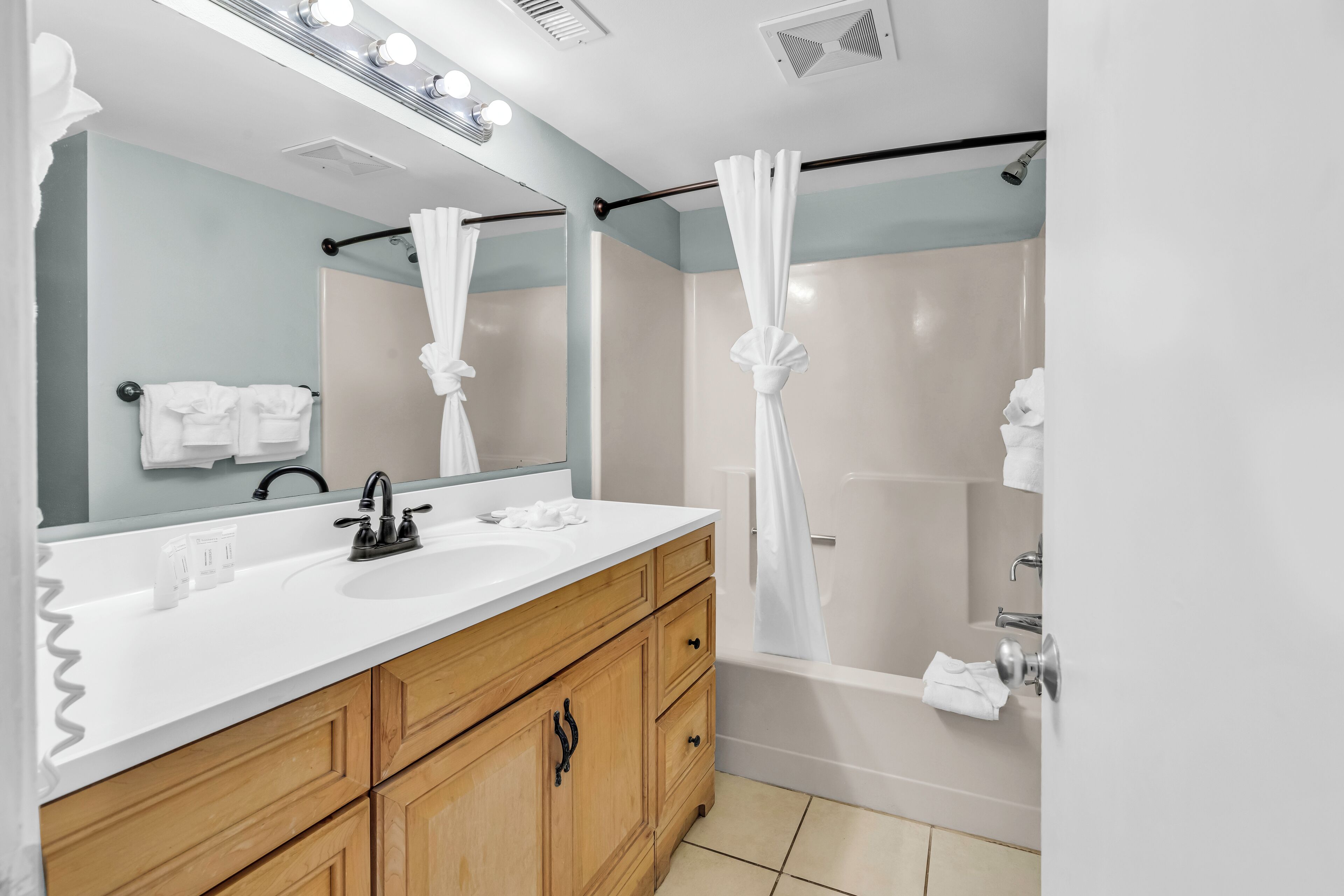 Un baño con lavamanos blanco, tocador de madera, un espejo grande y una ducha con cortina.