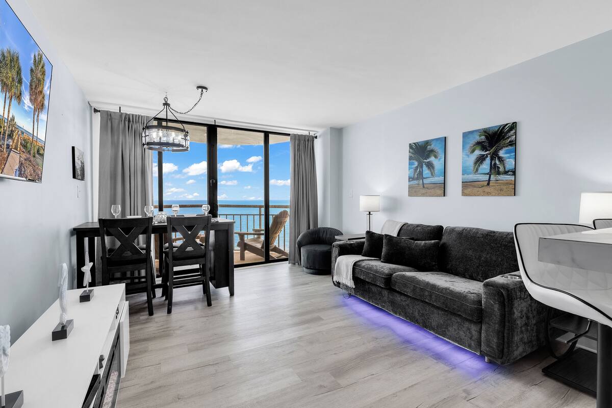 Una sala de estar moderna con un sofá, una mesa de comedor y sillas, un balcón con vistas al mar, y obras de arte en las paredes.